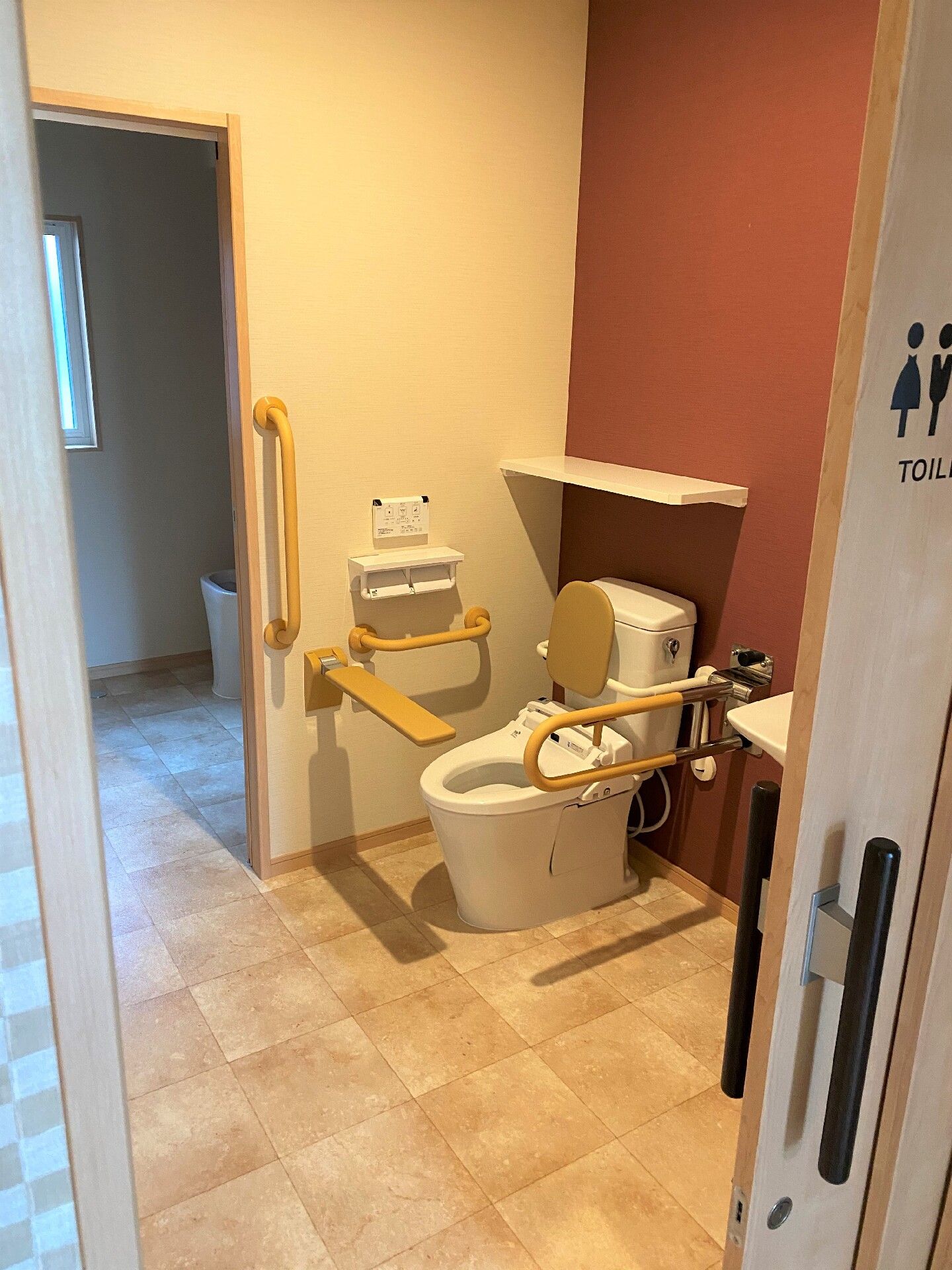 背もたれや手すり、前方にバーが付いた広いトイレ。手すりは両サイドに設置されていて、トイレのドアにはイラストも描かれている。