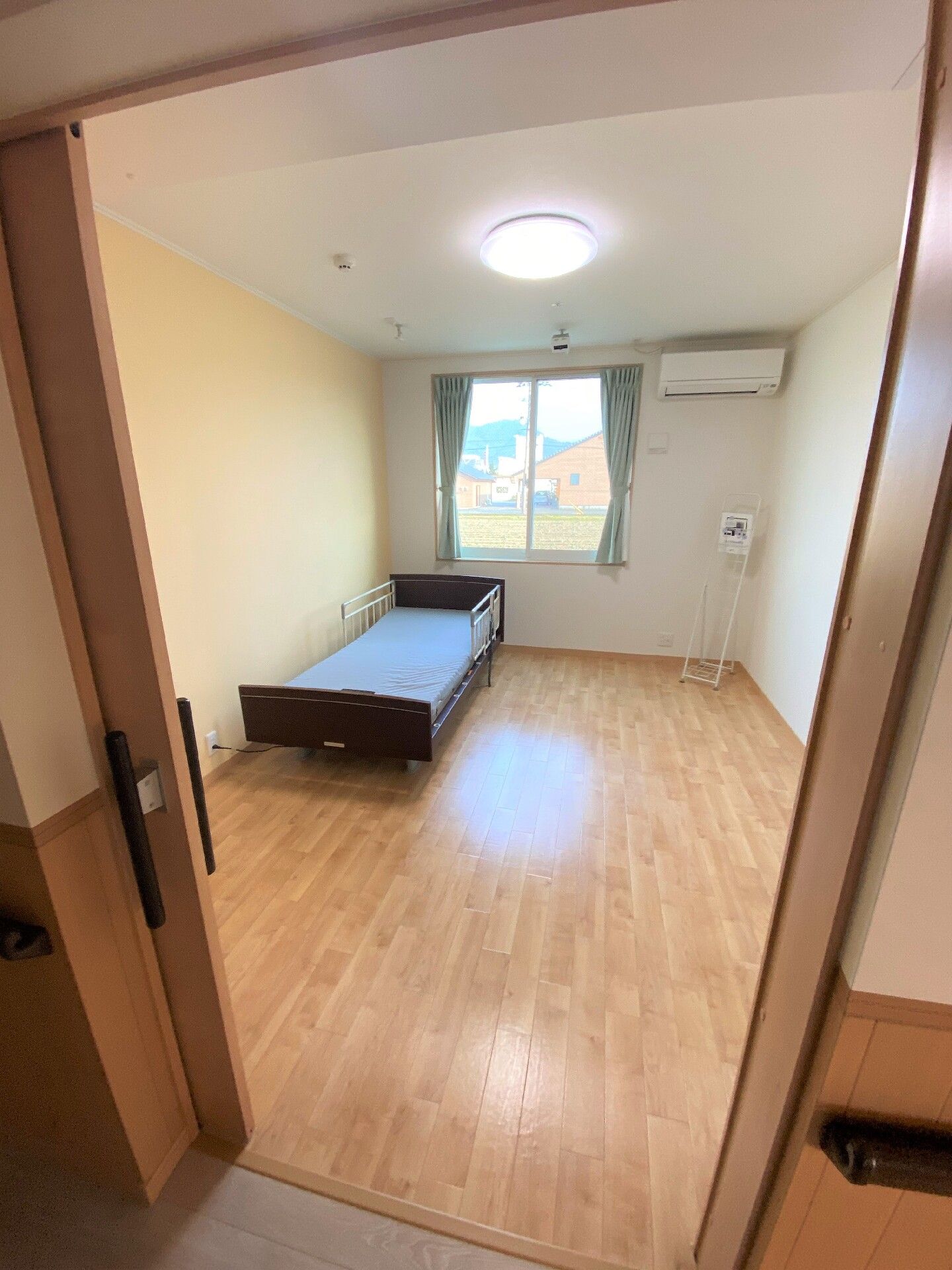 大きな窓にはカーテンが付き、壁にはエアコンが設置され、壁際には介護ベッドが置かれている。出入り口はスライド式のドアで廊下には手すりもある。