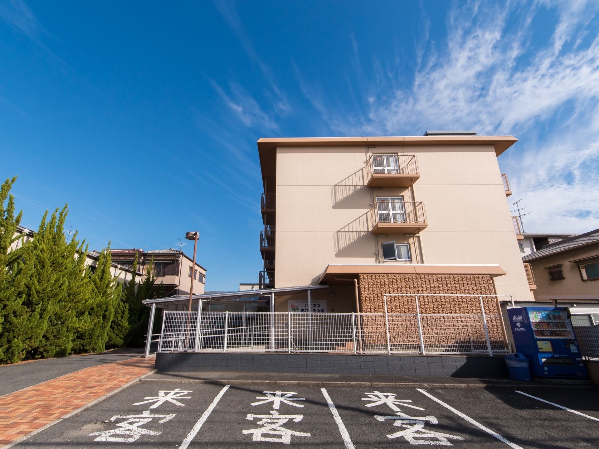 青空が広がる中に、コンクリート造りの4階建ての建物がある。建物横には来客用の駐車スペースが確保されている。
