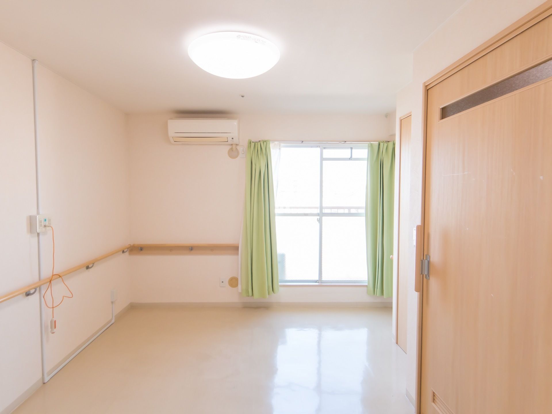 緑のカーテンがかけられた窓が部屋の奥にある。壁には、木製の手すりが取り付けてあり、ナースコールも設置されている。