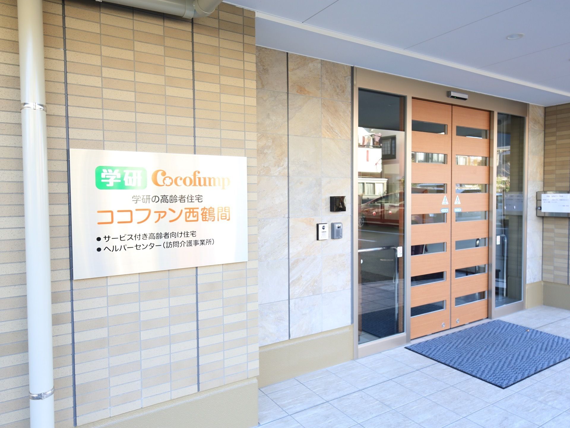 茶色い自動ドアが設置されており、両脇はガラスになっている。ブラウンのタイル風のサイディングの壁に金色の看板がかかっている。