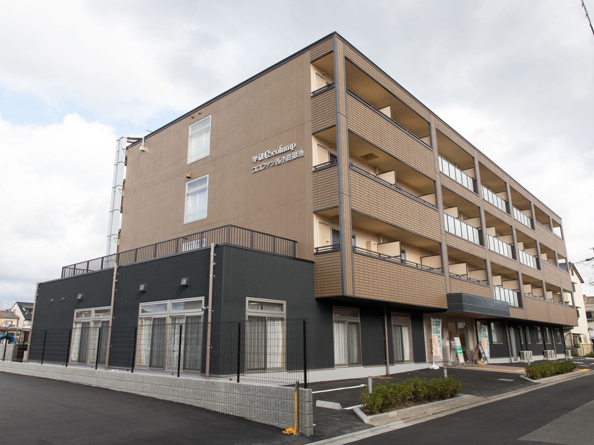 茶色と濃いグレーの外壁のモダンなマンション風の建物が建っている。建物の前には駐車スペースがあり、敷地と道路の境目付近に木が植えられている。