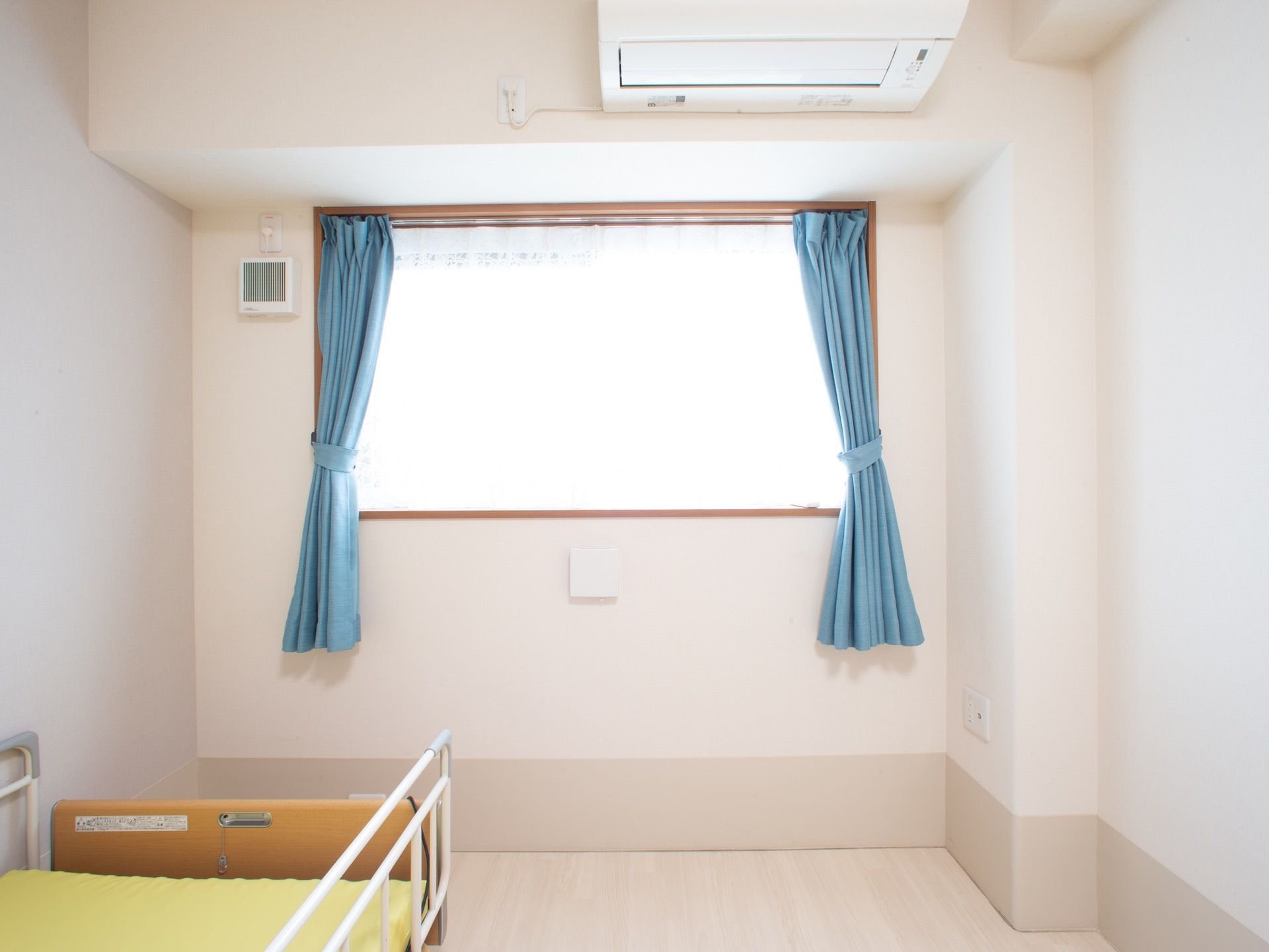 白い壁の居室にベッドが１台置かれている。青いカーテンがかかった窓があり、その上にはエアコンが設置されている。