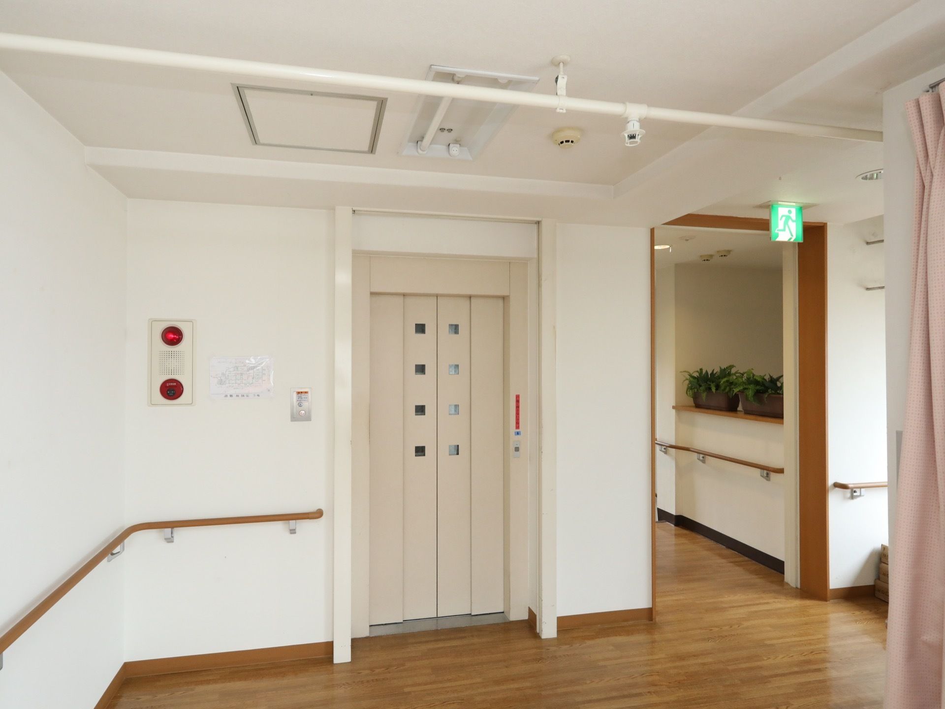 エレベーターの乗降口は、４つの四角形のスリットがついた白い扉になっている。左側の壁に非常ベルが設置されている。