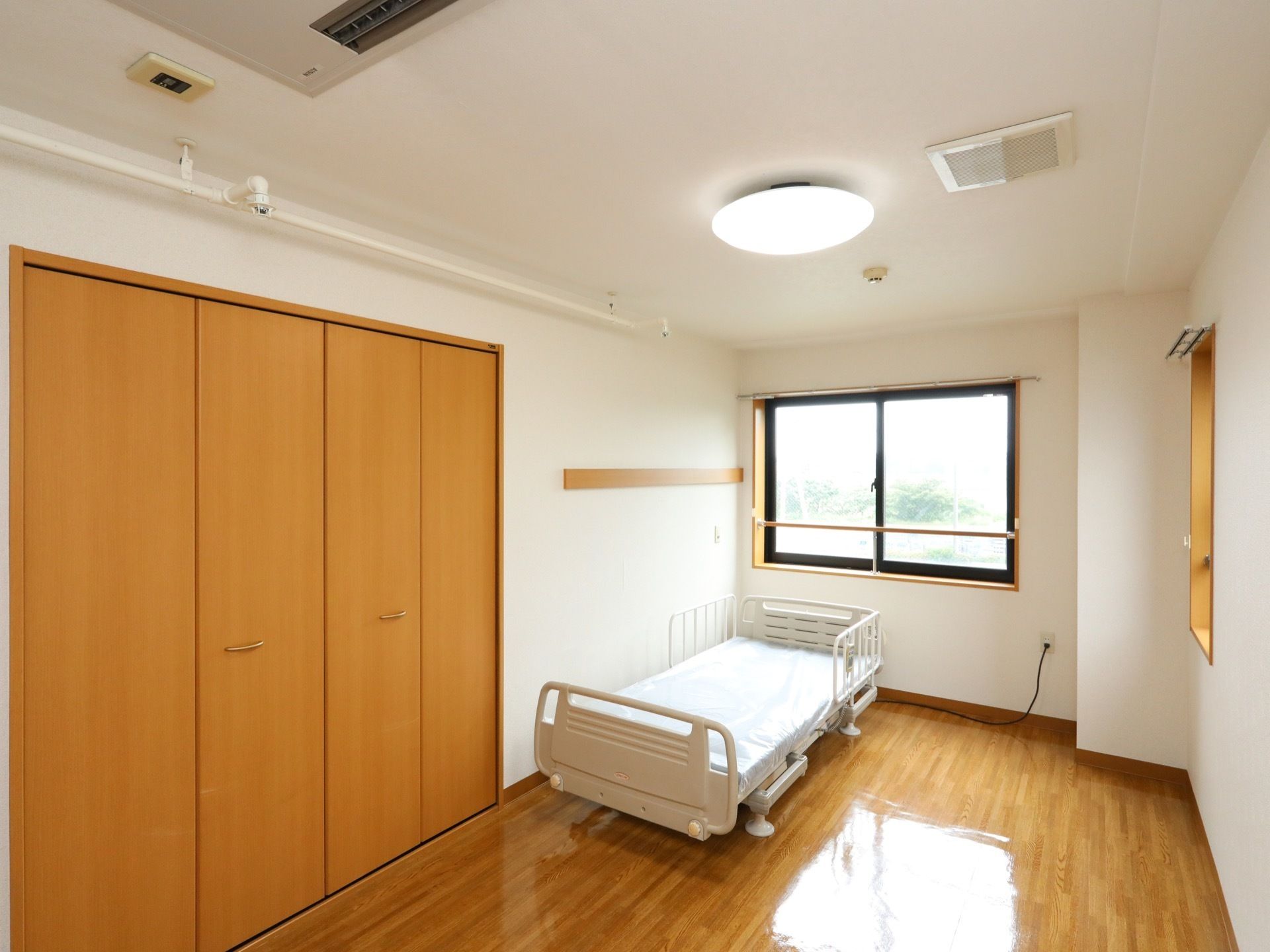 居室の窓際に、転落防止柵がついた介護ベッドが設置されている。天井近くまで高さのある扉がついた収納スペースがある。