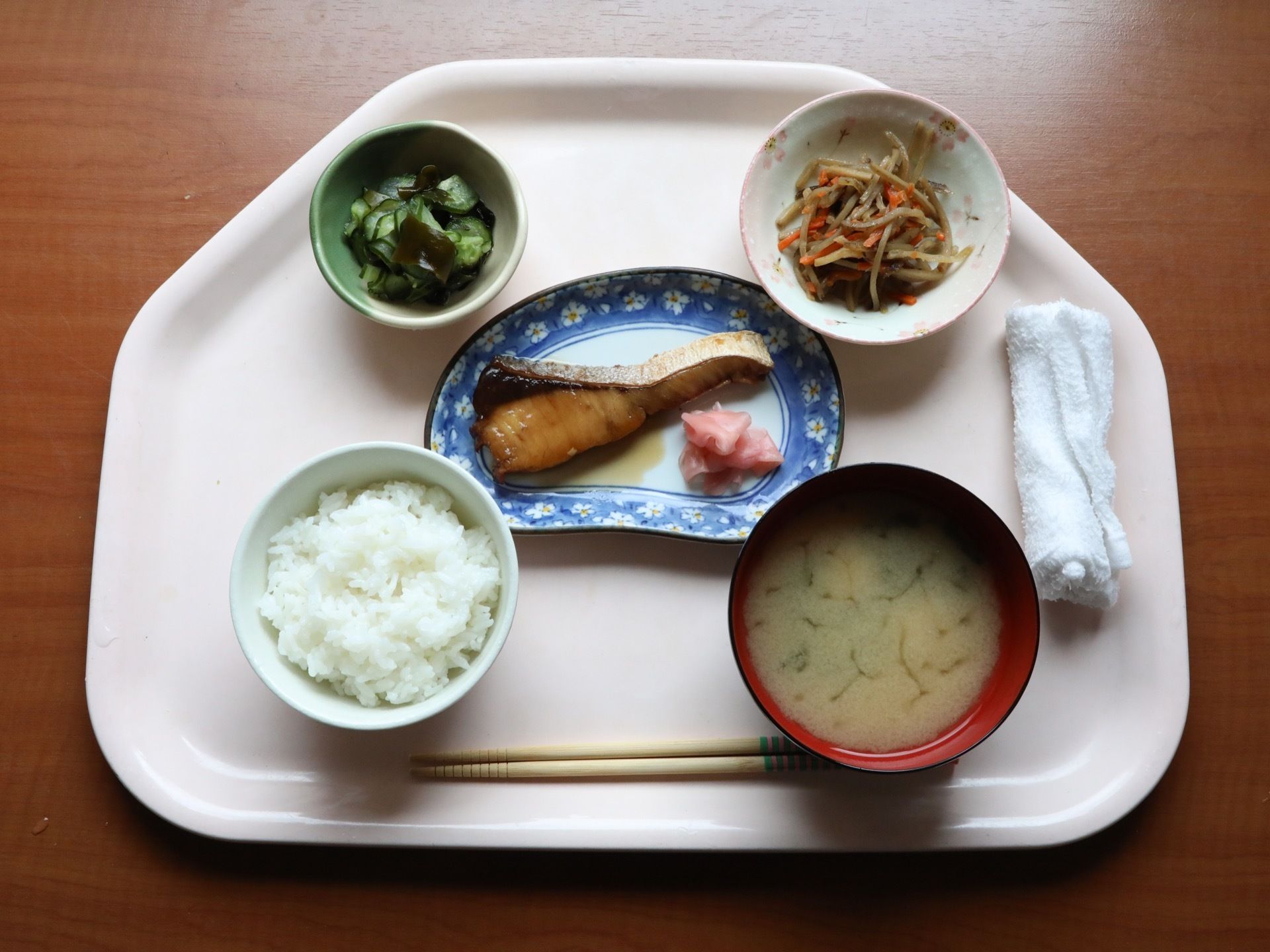 薄いピンク色のトレイに、ご飯、みそ汁、魚の煮つけ、酢の物などが載っている。おしぼりが丸めて置かれている。