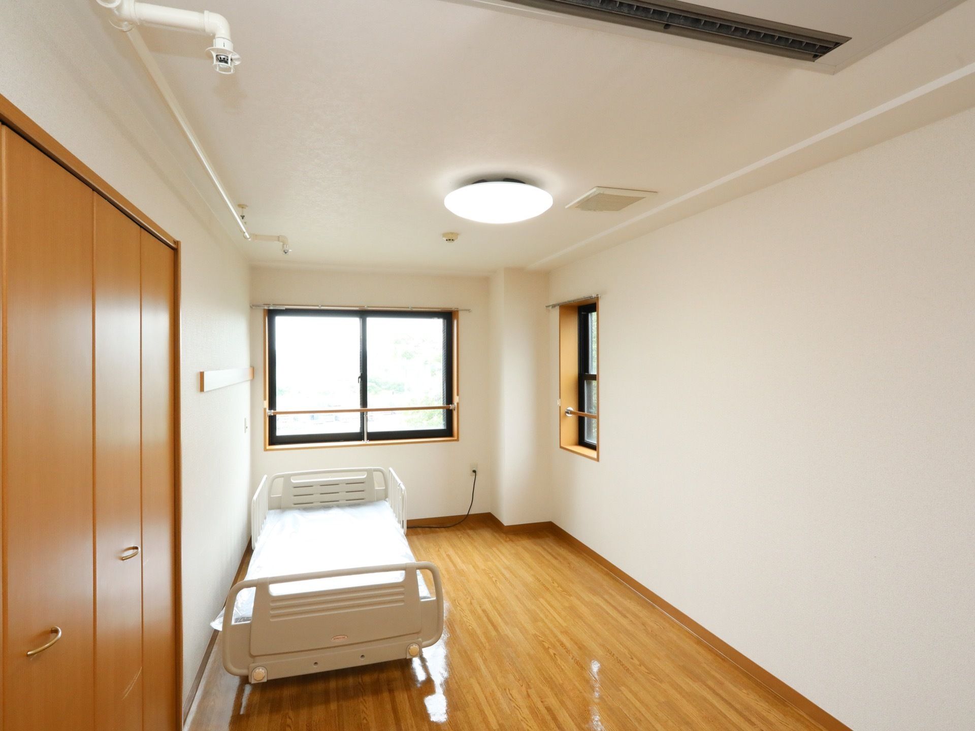 ２面の壁に窓がある居室である。転落防止柵がついた介護ベッドが壁際に設置され、その近くに収納スペースの扉がある。