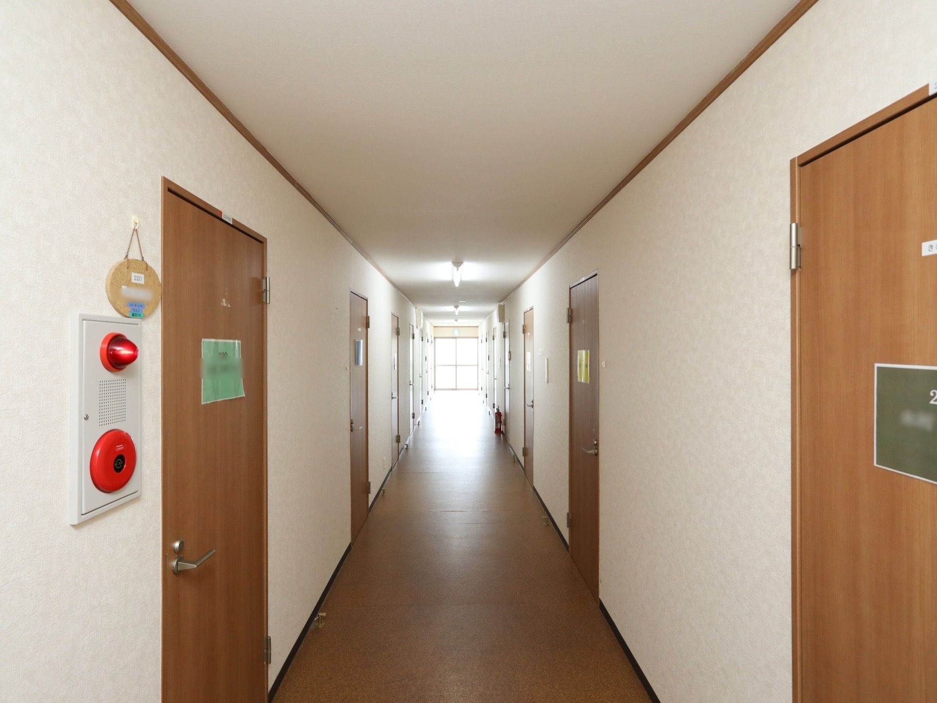 白を基調とした廊下で、両側に居室の入り口ドアがある。扉にはネームプレートが付いている。左手前壁に、火災報知器が設置してある。