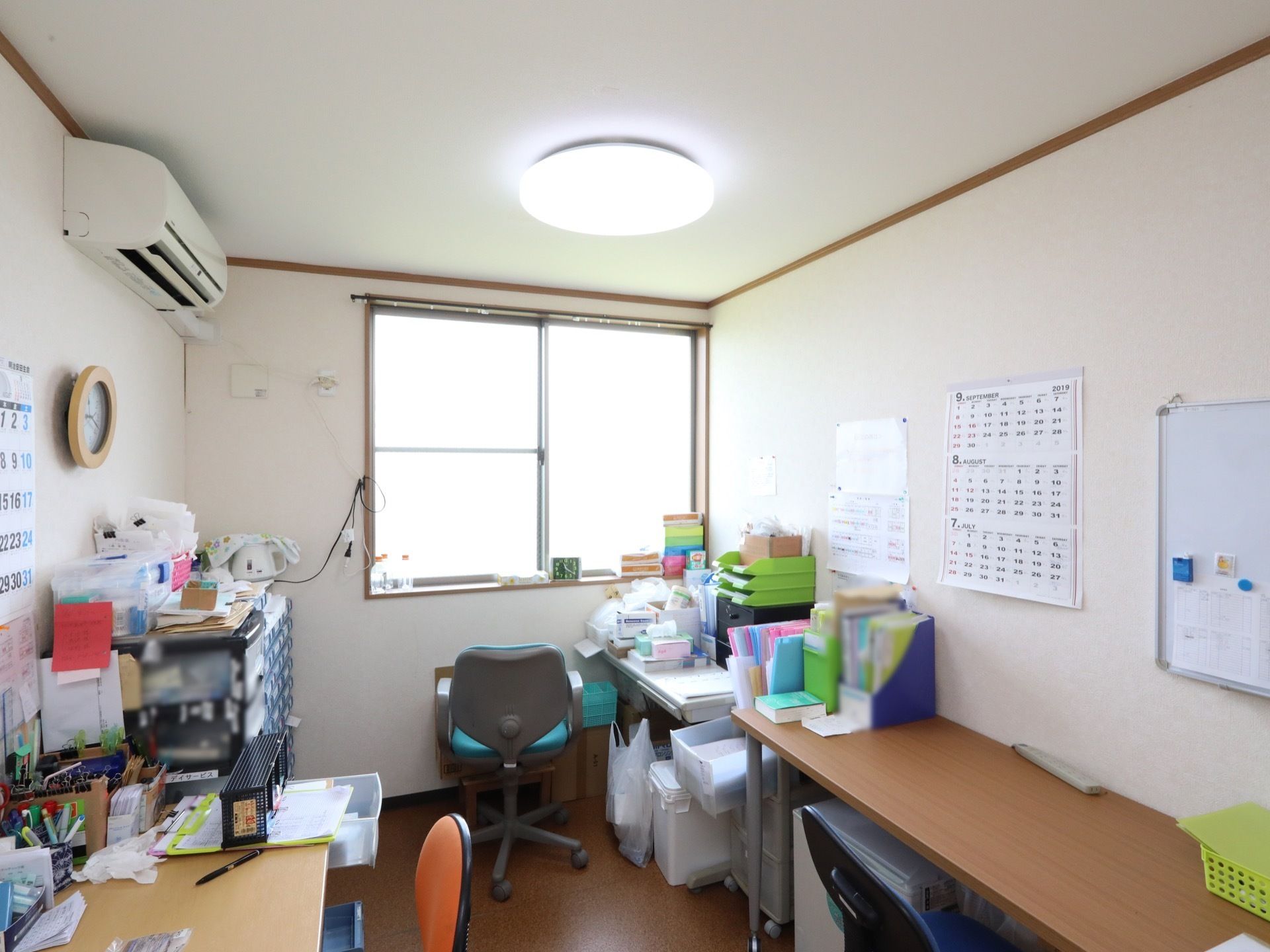中央に腰高窓があり、両側の壁に向かって、事務机が並んでいる。天井には丸型の照明が付けられている。机上には、多種の書類が置かれている。