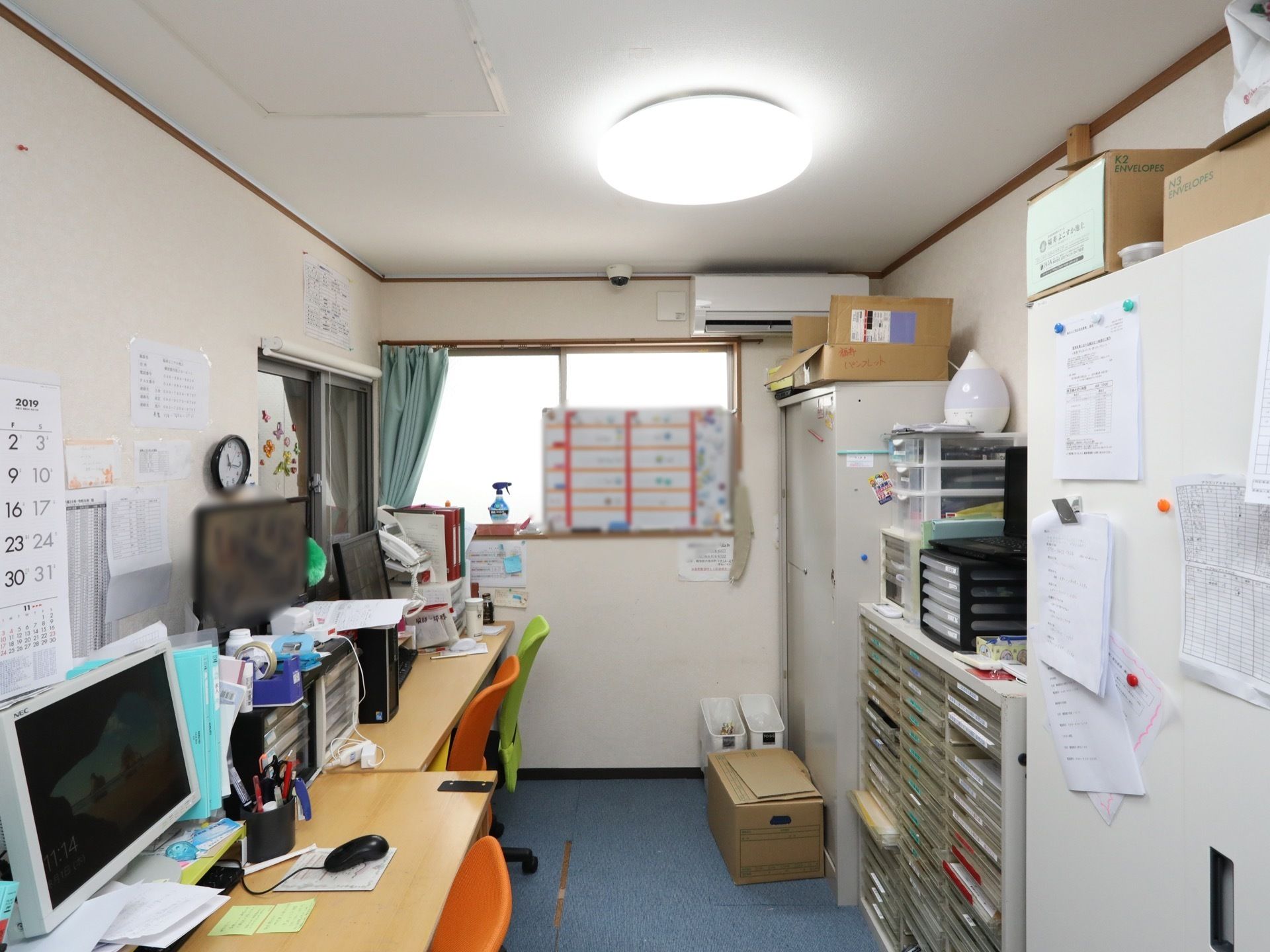 事務机があり、パソコンなどが置かれている。書類が保存されているキャビネットが並び、扉には伝達事項などの紙が貼り出されている。