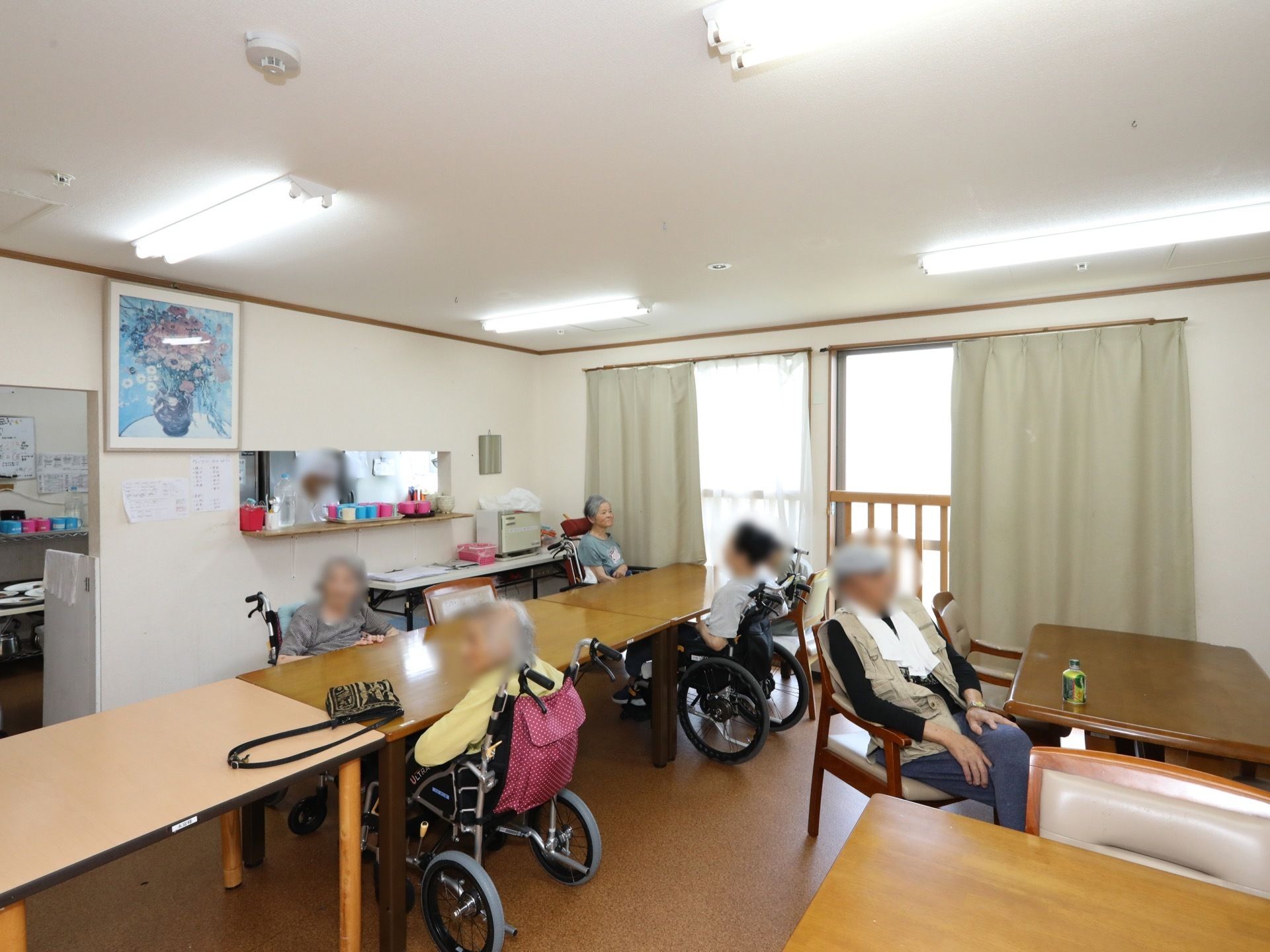 細長のテーブルが置かれ、数名の入居者様がくつろいでおり、車椅子を使用されている方もいる。壁に絵画が飾られている。