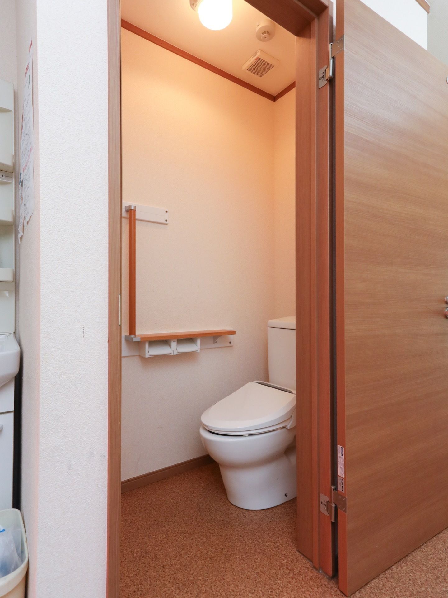 トイレの入り口に段差は無い。便座横の壁に、Ｌ字型の手すりが取り付けられている。天井に、火災感知器が設置されている。