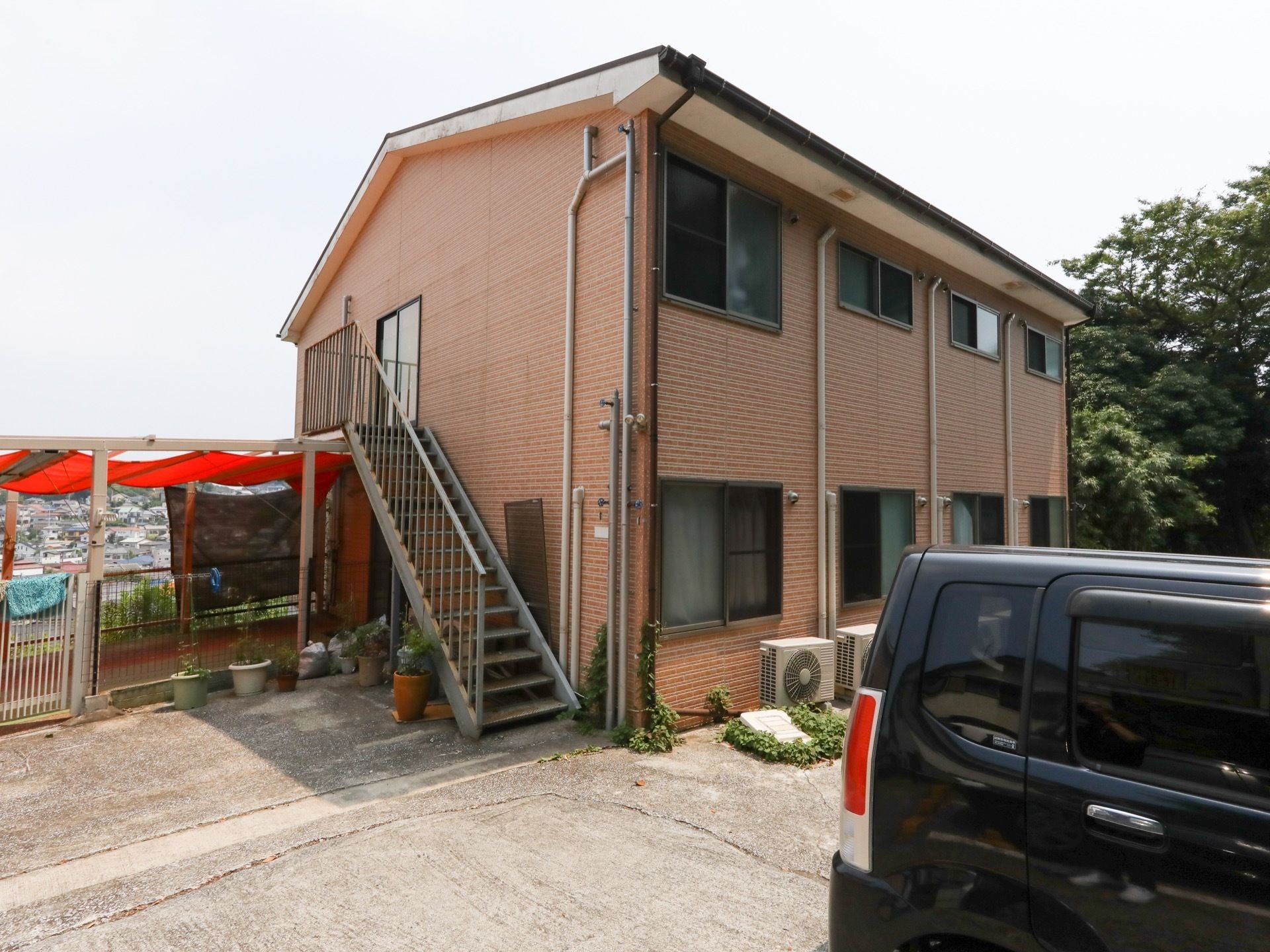 木造２階建ての建物で、外階段が設置されている。階段下に、草花の鉢が置かれている。建物の前に舗装がされた駐車場がある。