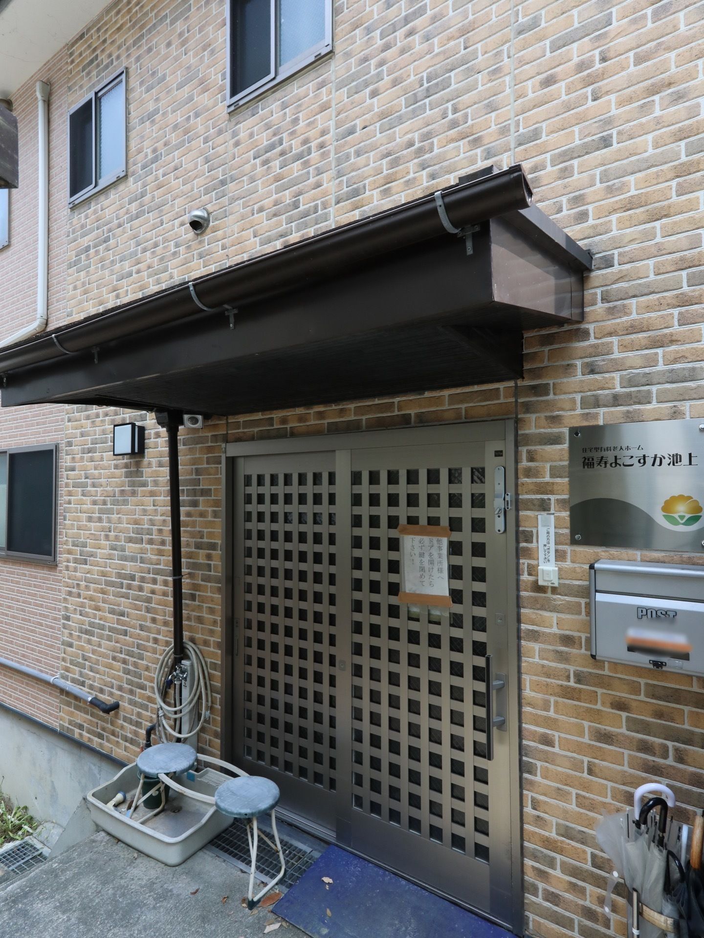 ひさしのある玄関。横には水道があり、ホースが巻かれている。右横の壁には施設名の入ったプレートが取り付けてある。