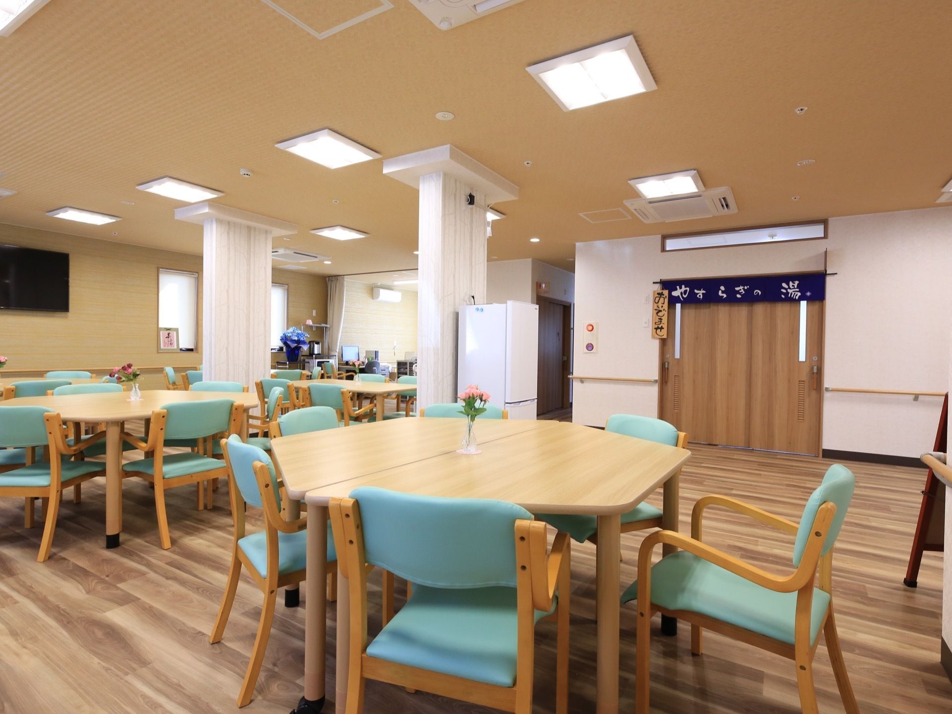 食堂に多くのテーブル、椅子がある。柱のそばに白色の冷蔵庫が置かれている。浴室の入口がある。