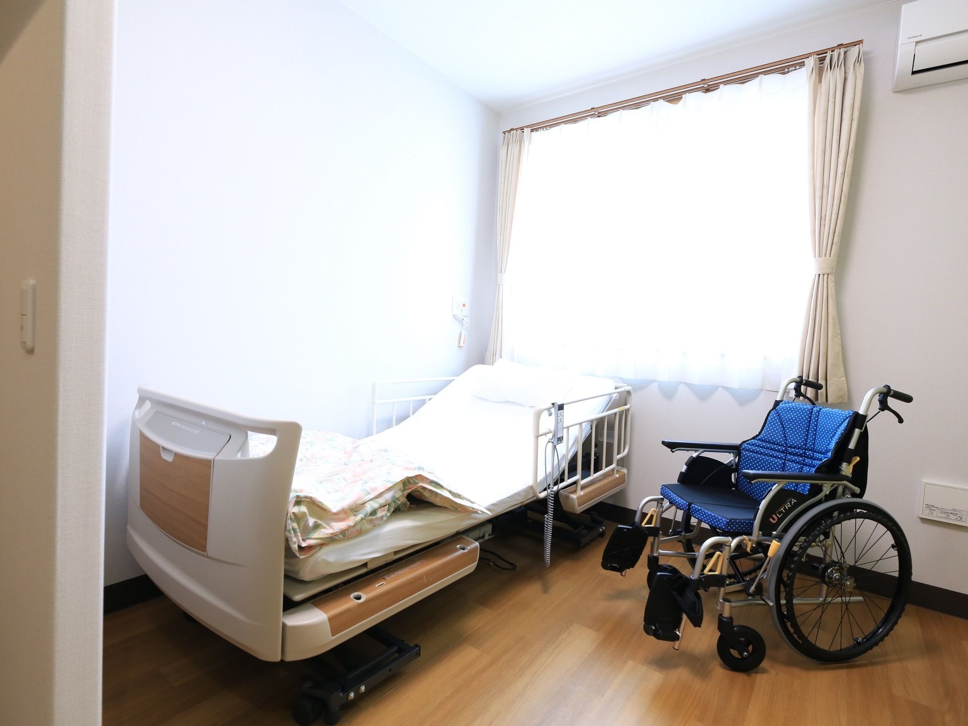 居室に横長の腰高の窓があり、レースのカーテンが閉められている。介護ベッドのすぐ横に車椅子が置かれている。
