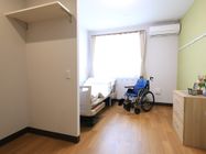 居室の窓の近くに介護ベッドが置かれ、窓側の方のパッドが持ち上がっている。窓の右上にエアコンが取り付けられている。