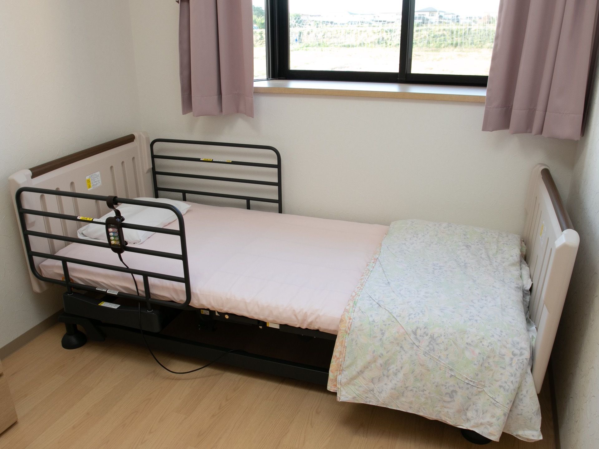 出窓が設置された居室に置かれている介護用ベッドである。落下防止のフェンスが装着され、リモコンが掛けられている。