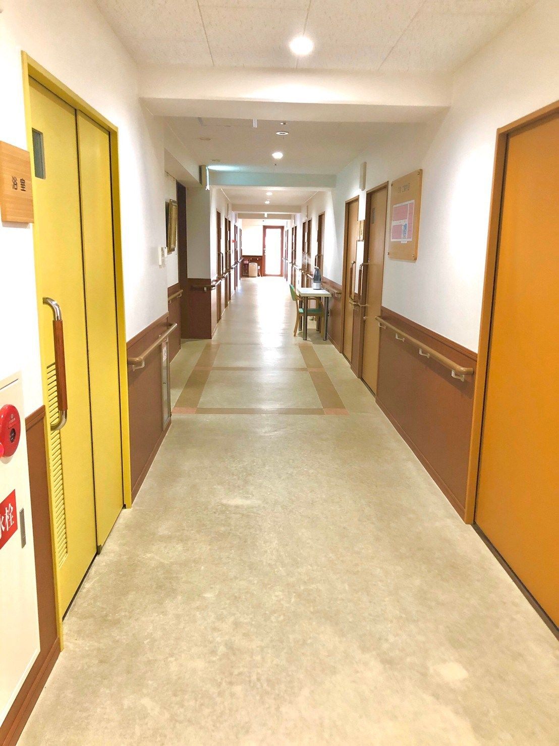 まっすぐな廊下には両側に手すりが付き、ダークブラウンの居室ドアが並び、左手には黄色いドアの共有スペースがある。