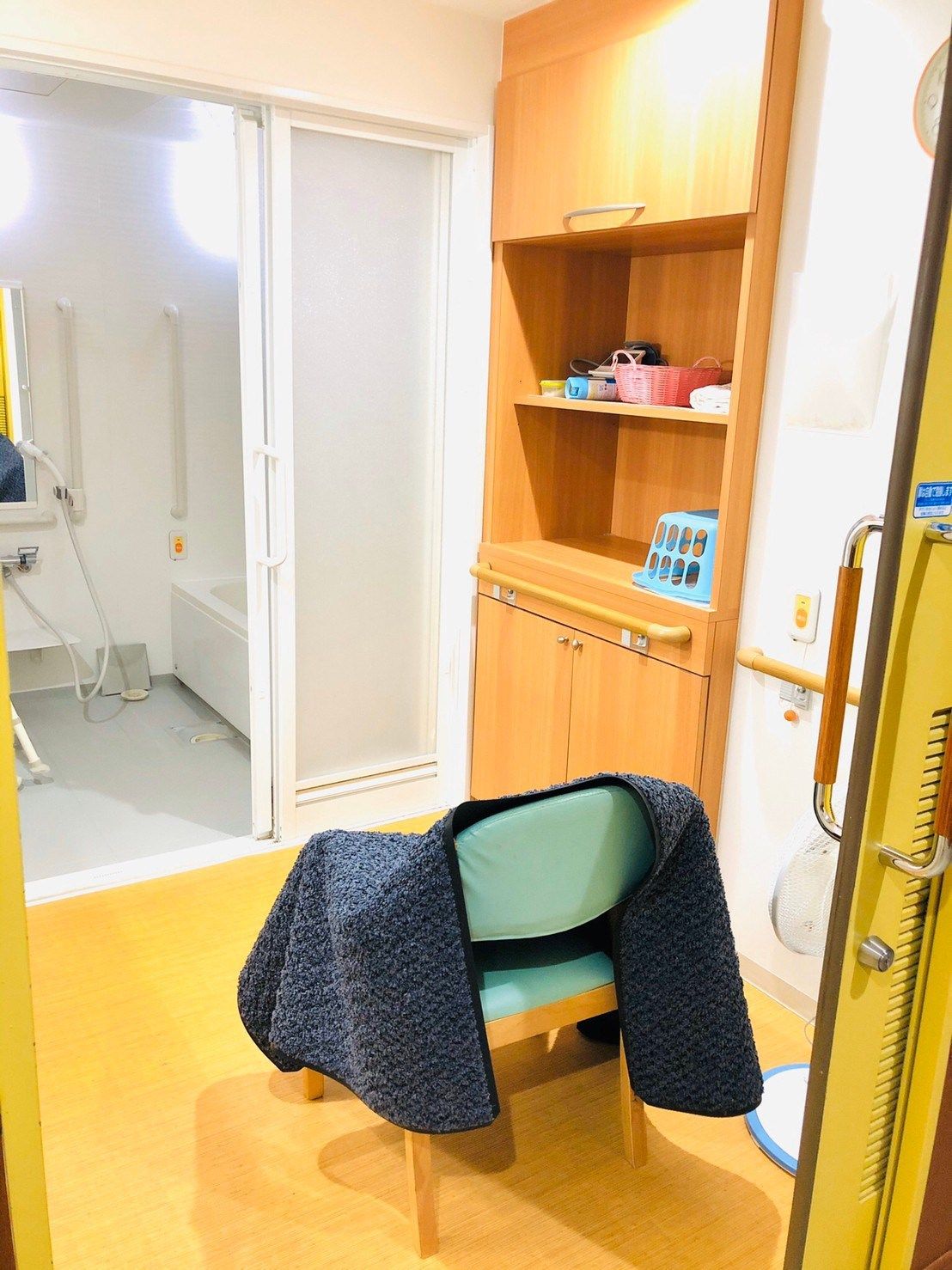 布がかけられた椅子が脱衣所の棚の前に置かれ、奥には白をメインとした浴室がある。