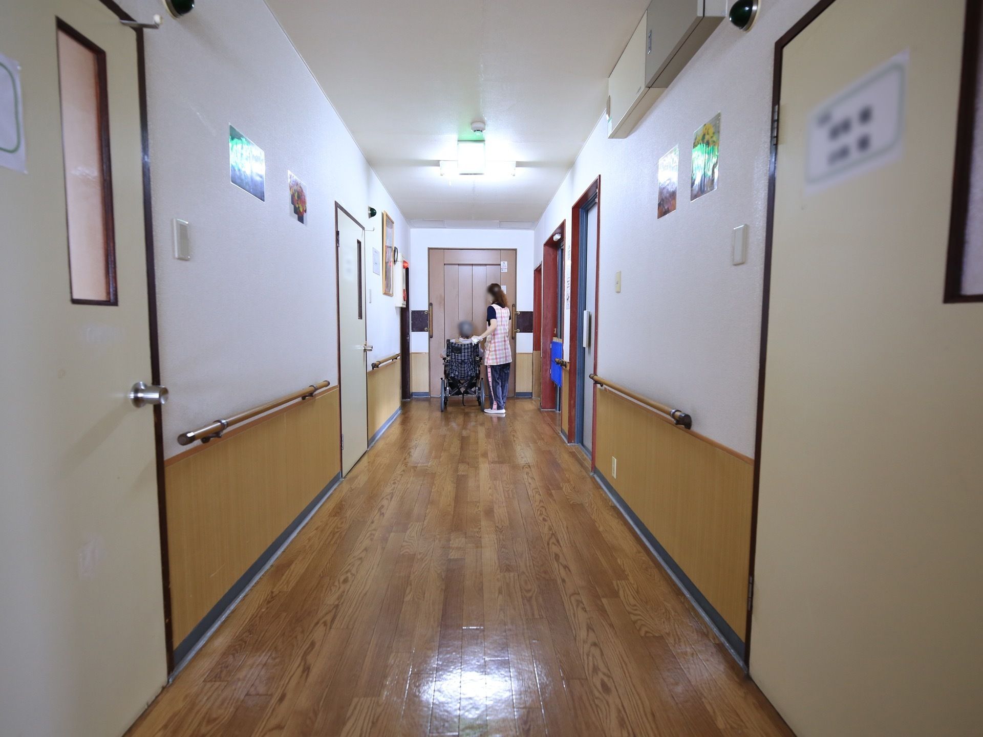 フローリングの廊下が伸びている。通路幅にはゆとりがある。車椅子に座った入居者様とスタッフが扉の前にいる。