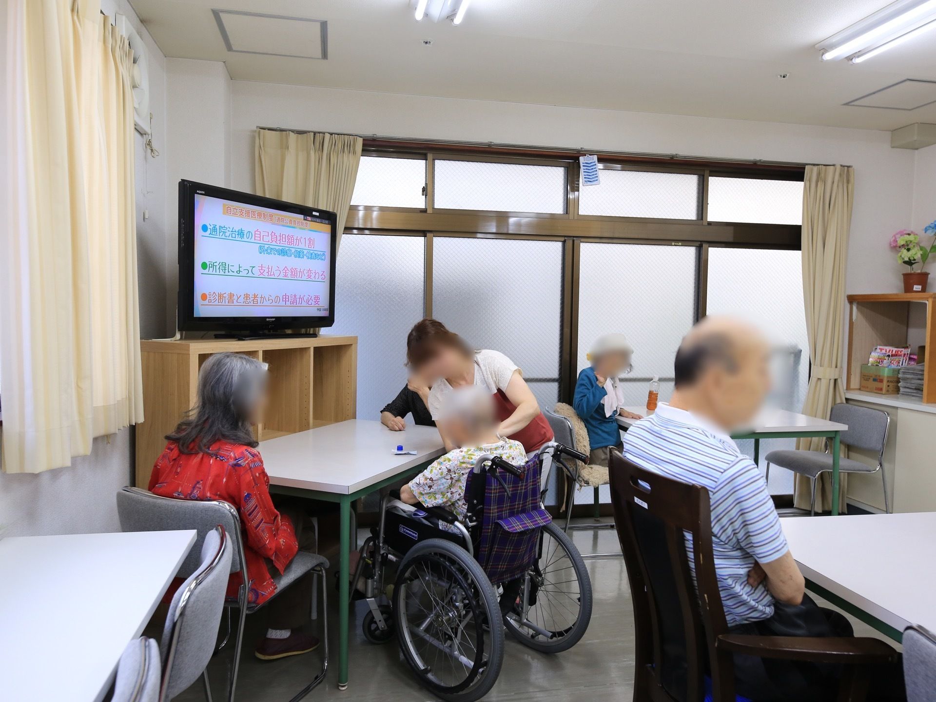 テーブルセットが４つほど置かれた部屋で、入居者様が数名腰かけている。テレビの近くではスタッフと談笑している方もいる。