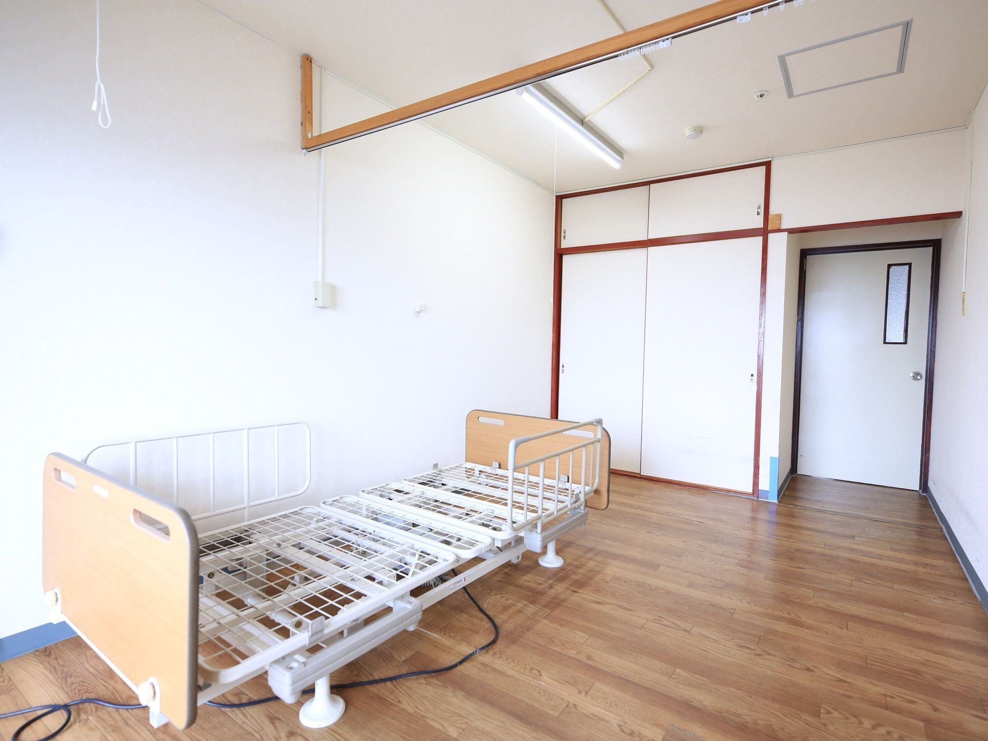 フローリングの居室に電動ベッドの骨組みが置かれている。ふすまタイプの収納があり、天井には照明器具が付いている。