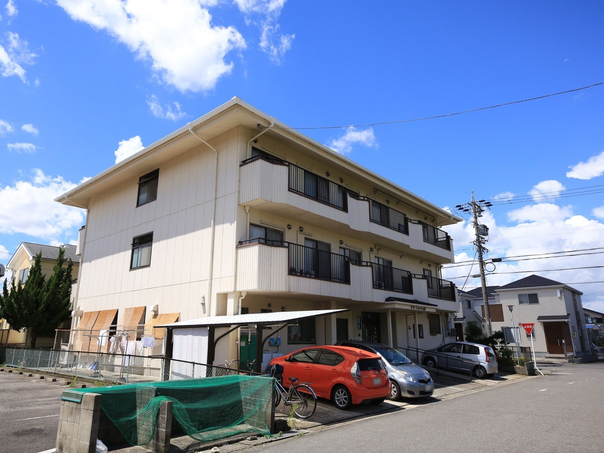 マンションタイプの3階建て住宅がある。建物の前は駐車場になっていて、車が3台止まっている。屋根の付いた駐輪スペースも設けられている。