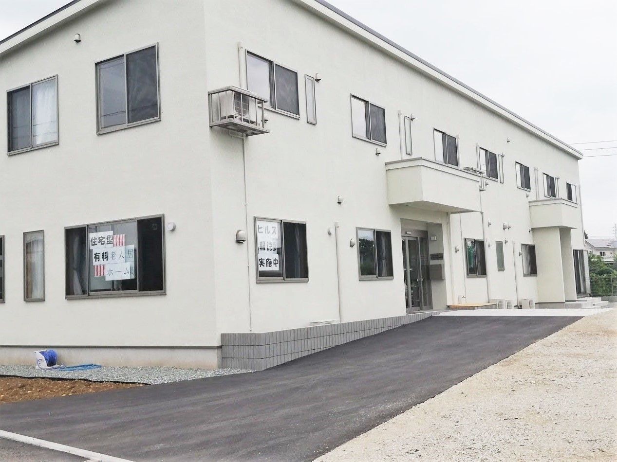 2階建ての建物は白をで屋根が付いた玄関があり、玄関前には塗装された道がある。