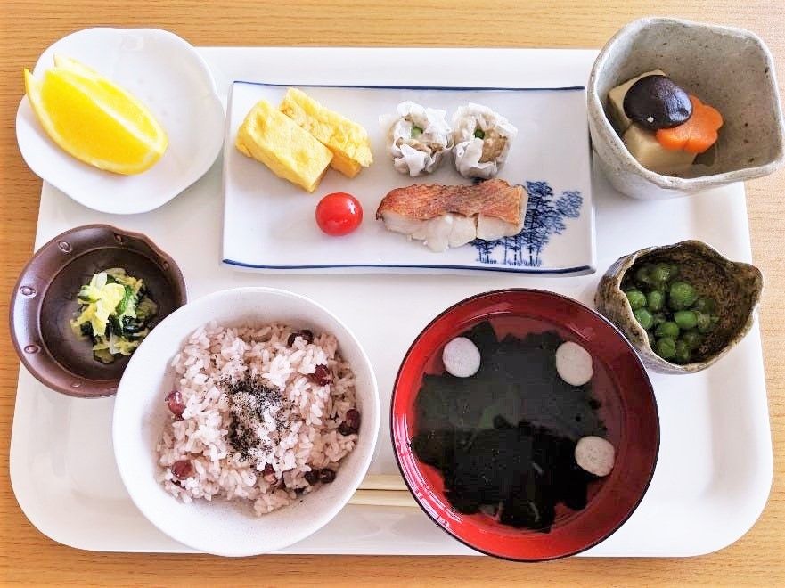 焼き魚やシュウマイが乗った皿を中央に、赤飯に汁物、フルーツや煮物など小鉢が並んだ白いトレー。
