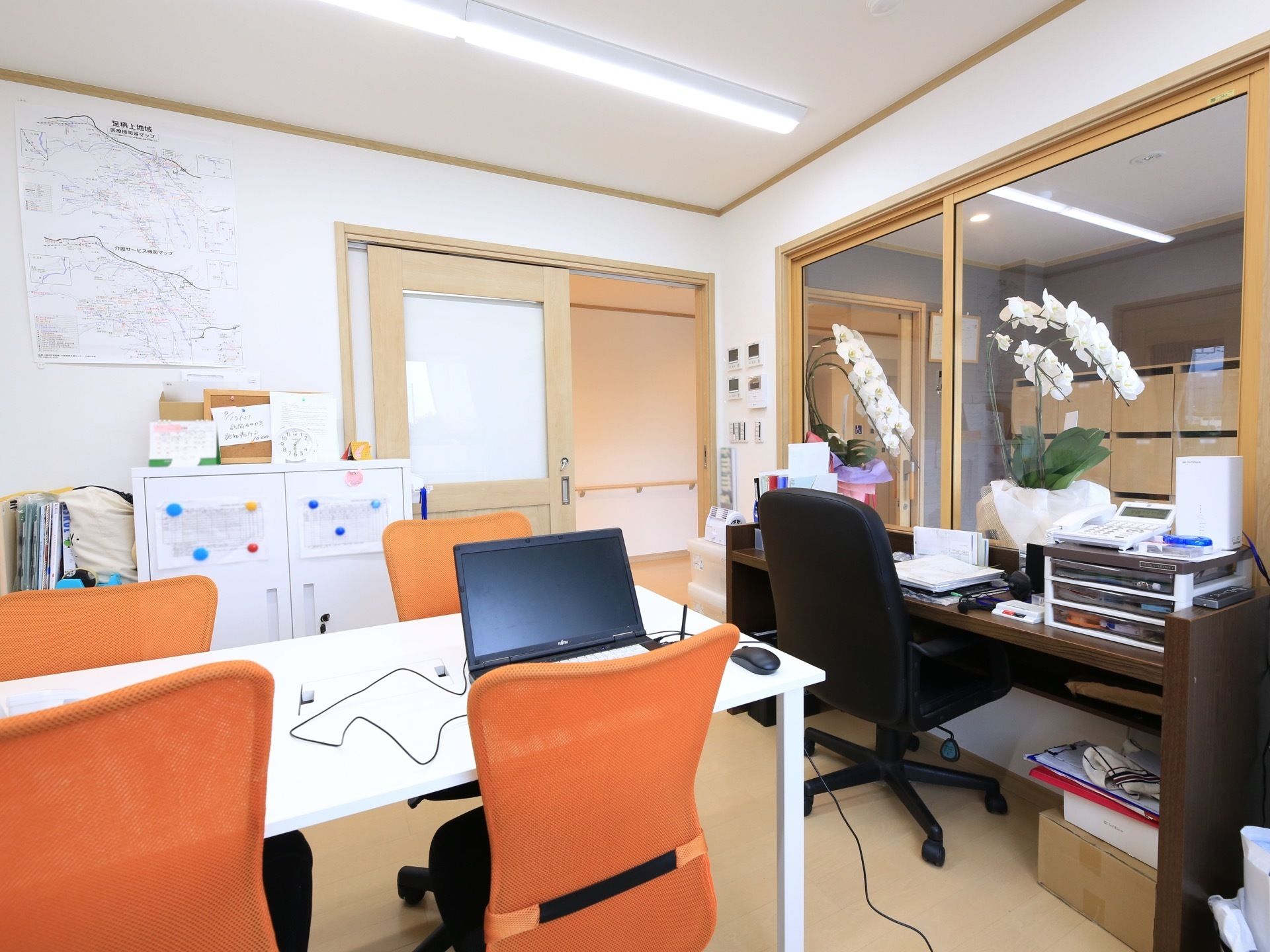 部屋には、白いテーブルにオレンジ色の椅子が複数置いてあり、テーブルの上にノートパソコンが1台ある。側面は、ガラス張りの受付窓口になっている。