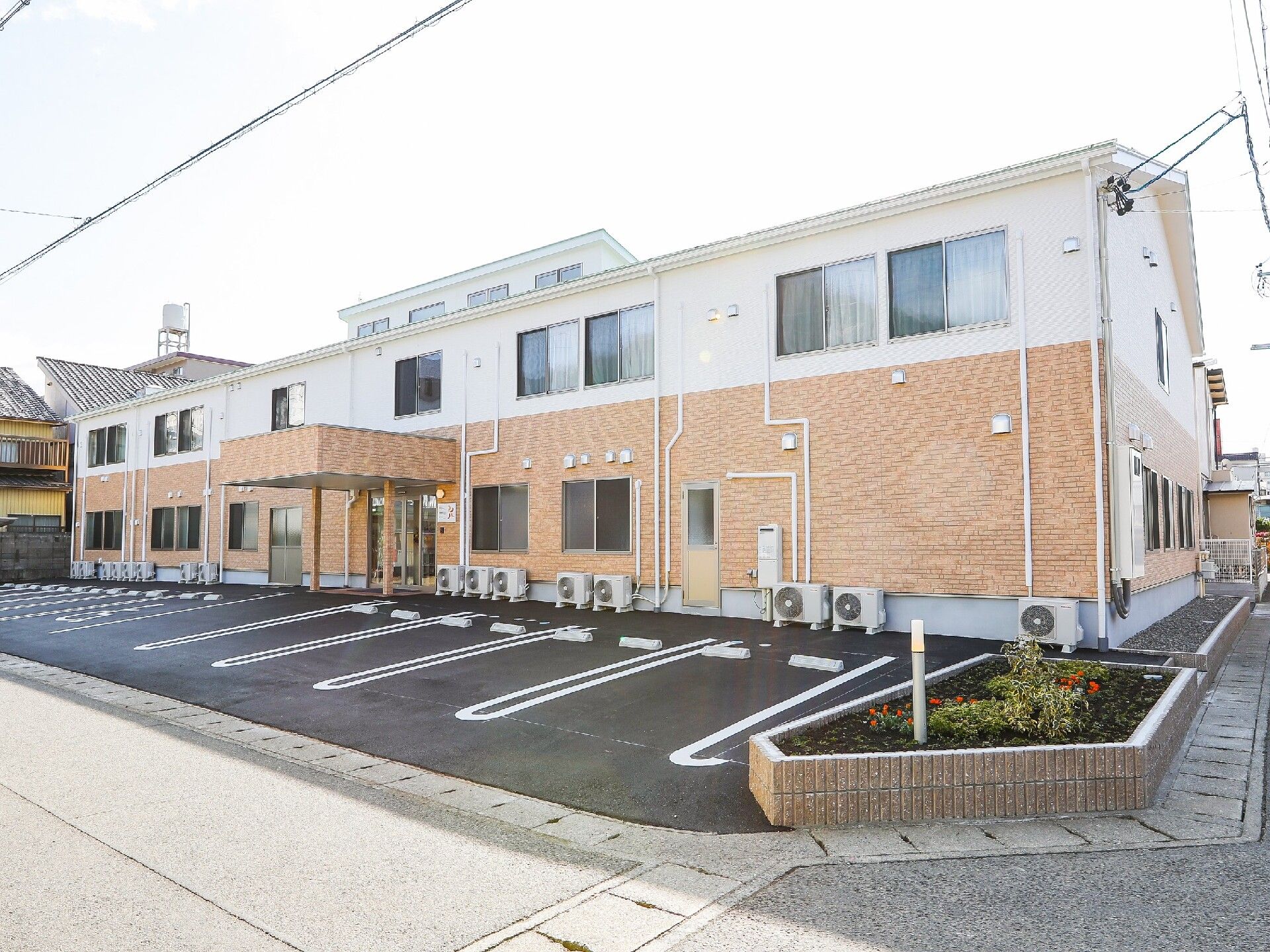 2階建ての建物は1階部分がベージュ、2階部分が白で、天窓も付いている。玄関には屋根が付き、建物の前には広い駐車スペースもある。