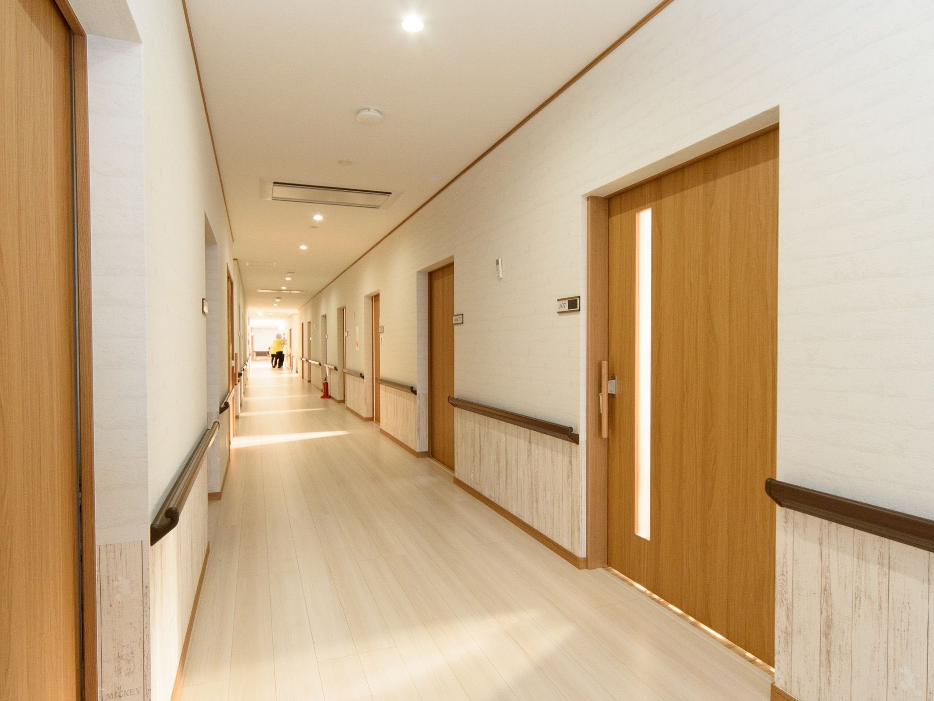 白と木目調がさわやかな印象である。広い廊下の両側に、居室の引き戸が並んでいる。廊下には手すりが設置してあり、安全に行き来できる。