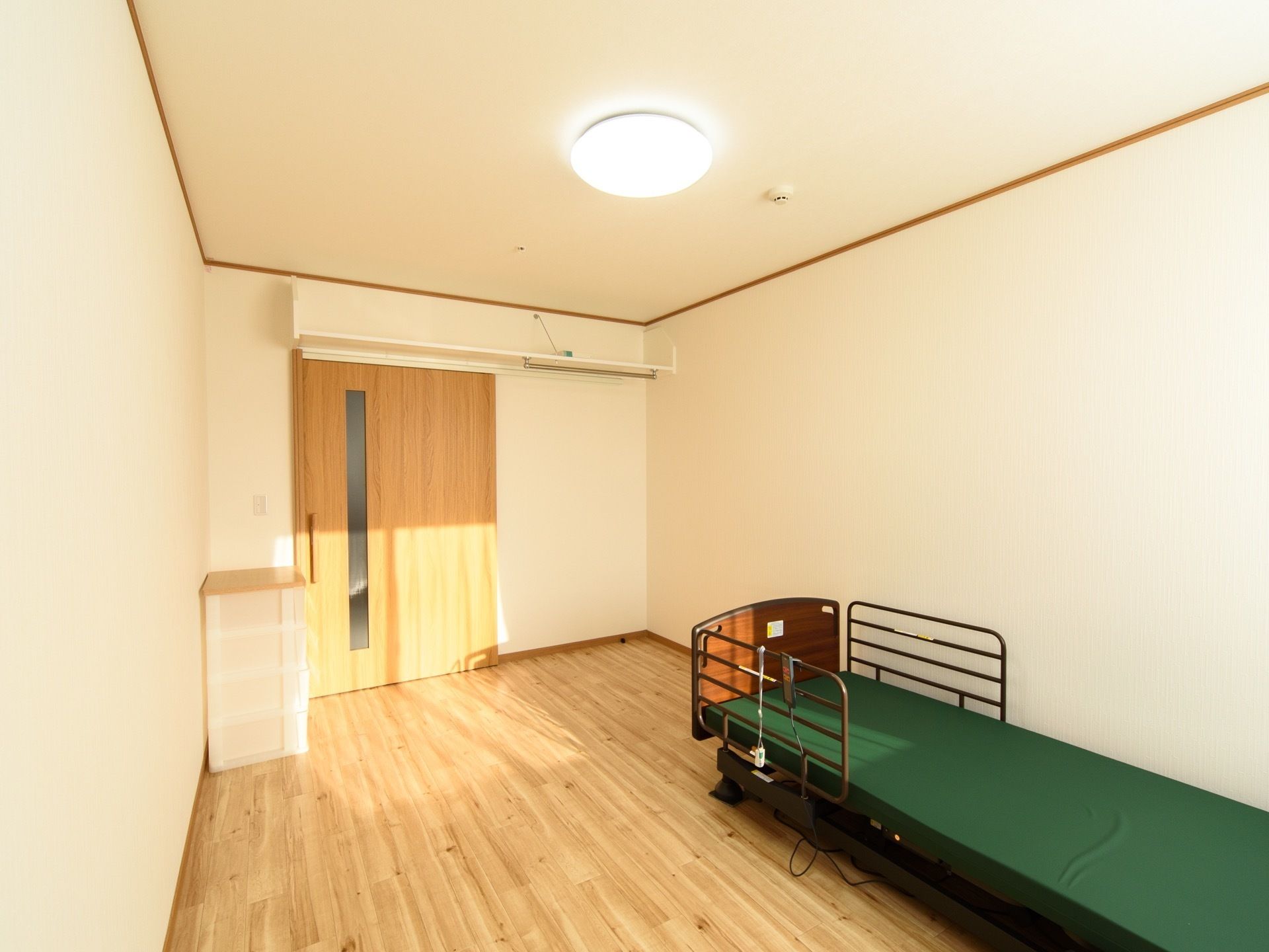 白壁、木調のフローリングの1人用居室である。介護用ベッドと衣装ケースがある。部屋はシンプルで清潔感があり、とても明るい。