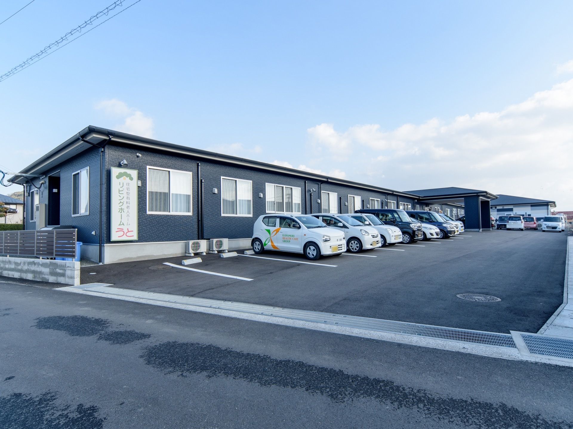 平屋建ての建物に看板が見える。建物前には駐車場があり、車をご利用の来訪者様にも便利である。施設周辺に高い建物がなく開放的だ。