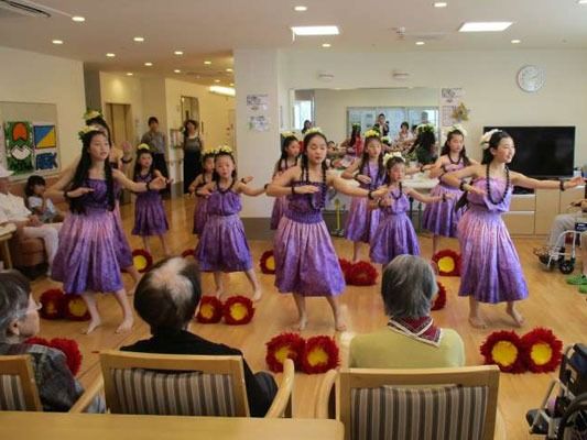 紫のドレスを着て優雅にフラダンスを踊る女の子たちの姿を椅子に座って眺める入居者。