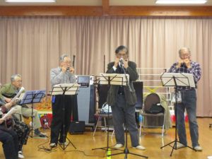 高齢男性がギターやハーモニカで演奏をしている様子。どの人も眼鏡をかけている。