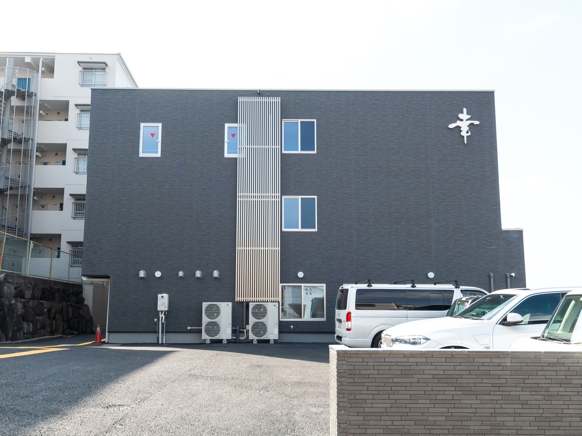 黒色の外壁で、三階建の建物である。建物前には駐車場があり、右側には車が停められている。建物の左側には集合住宅が見える。
