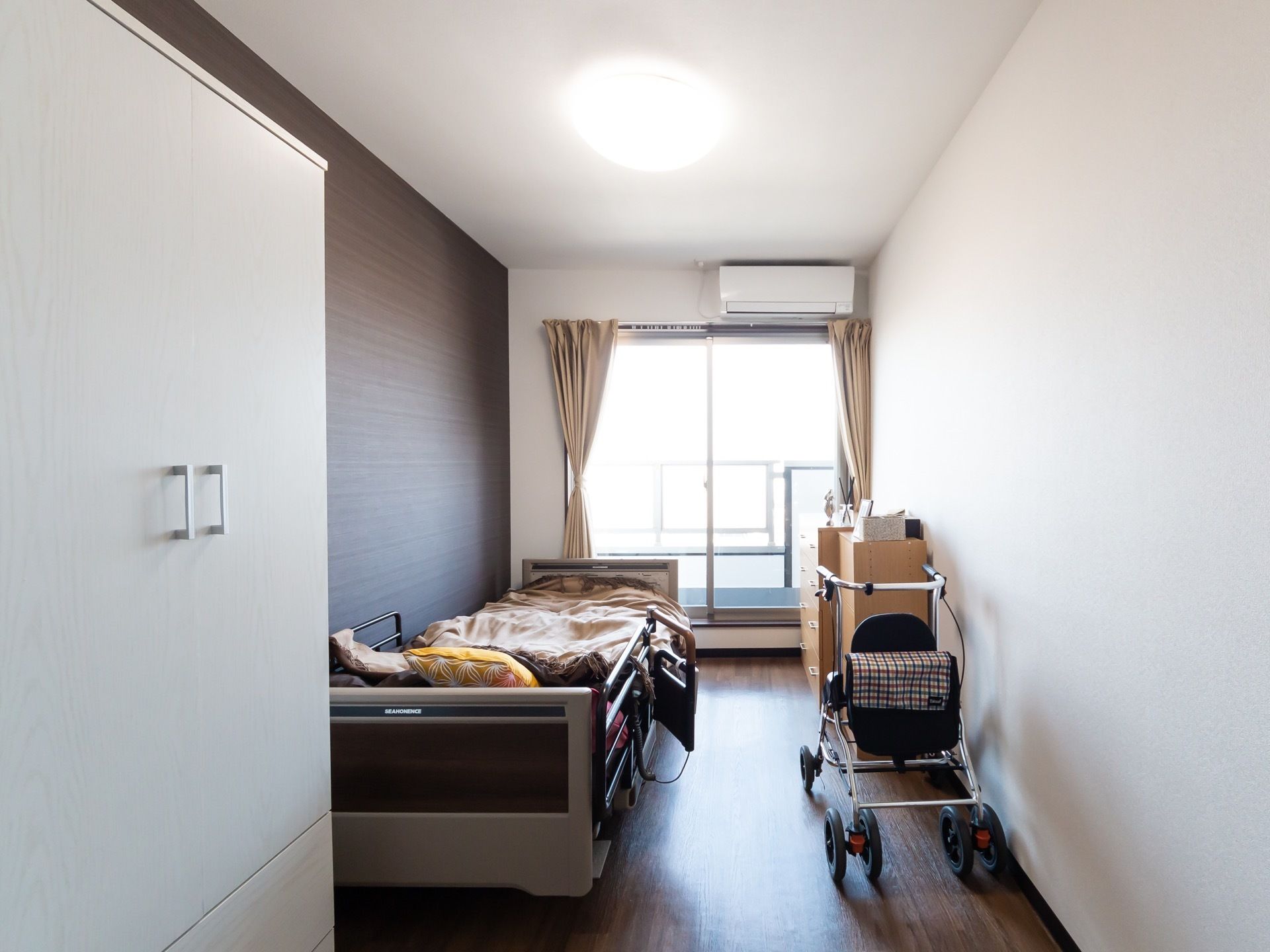 白色の壁に茶色の床の居室である。奥には窓があり、上部にはエアコンが取り付けられている。左側にはベッド、手前には白色の収納がある。