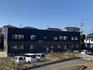黒い外壁の2階建ての建物の前には駐車場があり、数台の車が止められている。奥には住宅地も見える。
