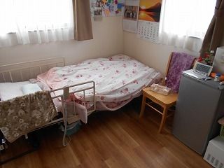 冷蔵庫やいす、寝具付きのベッドが置かれたフローリングの部屋には、カレンダーなども飾られている。