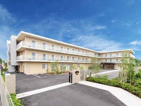 大きな駐車場が付いた3階建ての建物はベージュ系の外壁でエル字型。日当たりのいい場所に建っている。