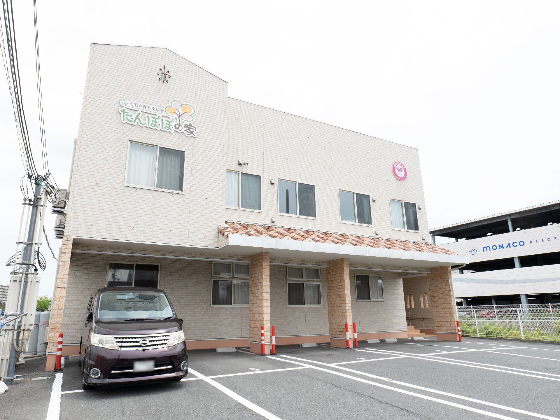 ベージュの建物は２階建てで外壁には住宅名も書かれている。駐車場付きで日当たりのいい場所に建っている。