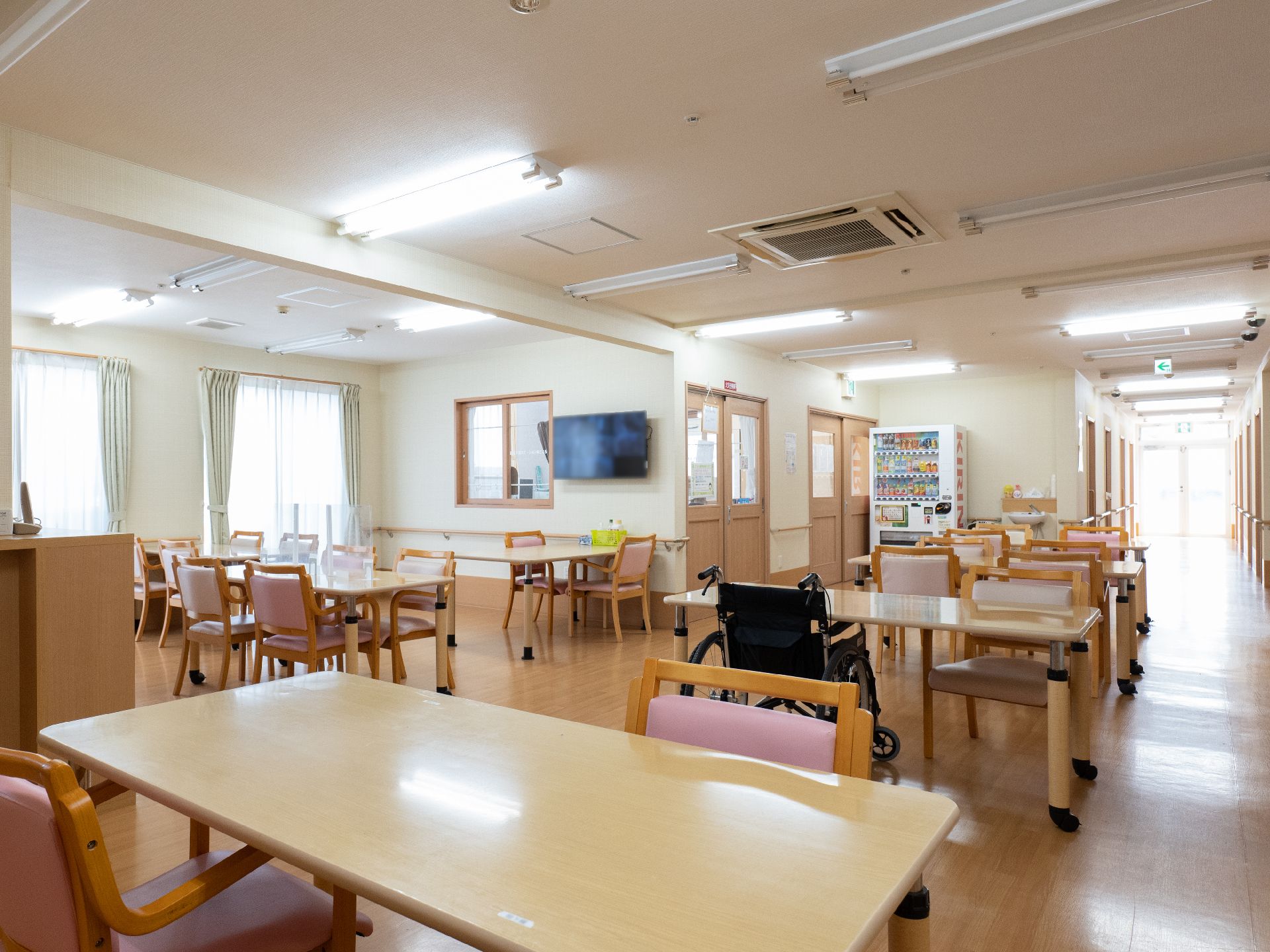 木製の机とピンクのクッションが付いた椅子が並んでいる白い壁の部屋