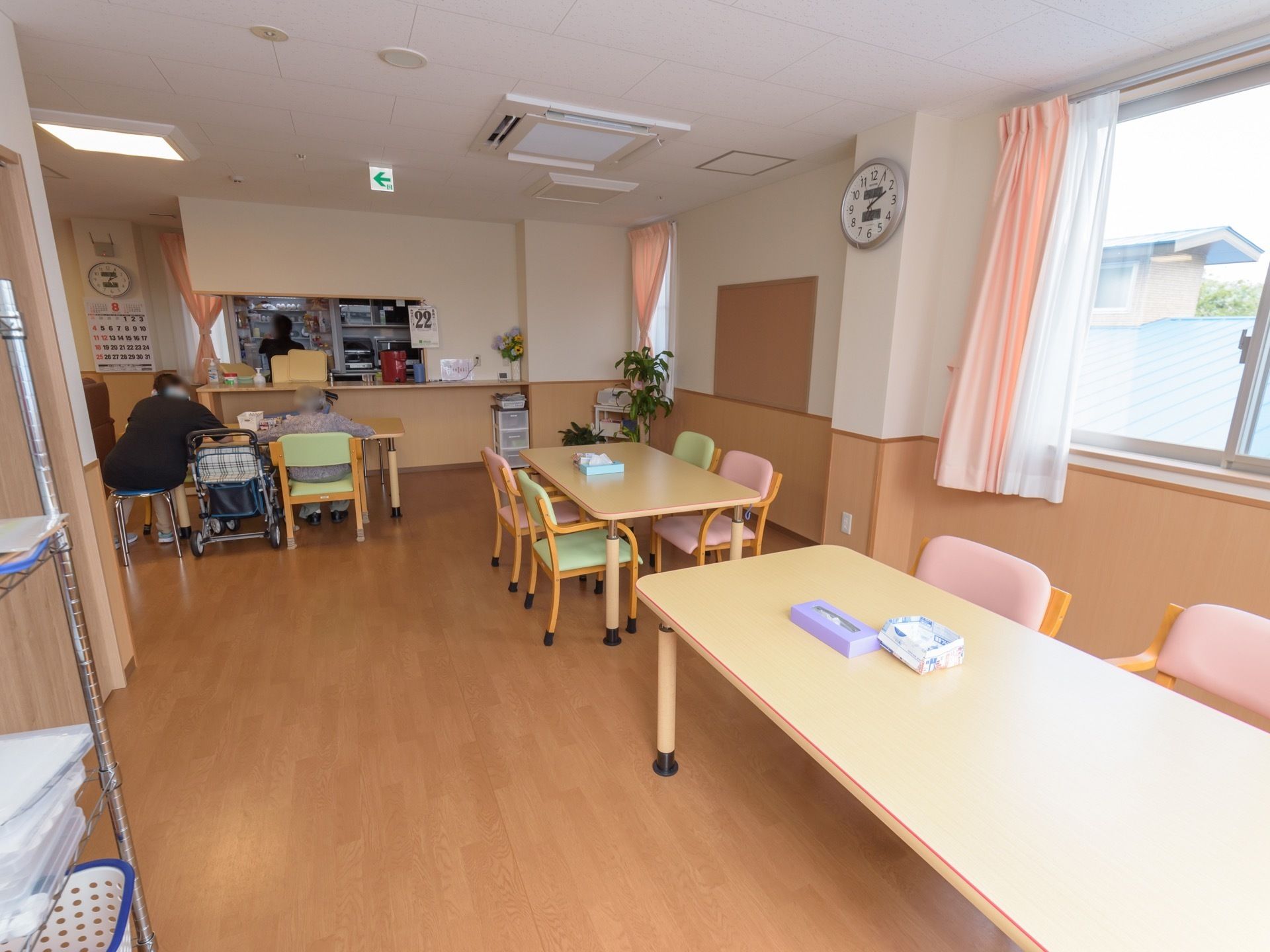 カウンターテーブルの手前に置かれたテーブルに２人が腰を掛けている。壁に時計やカレンダーがかかっている。