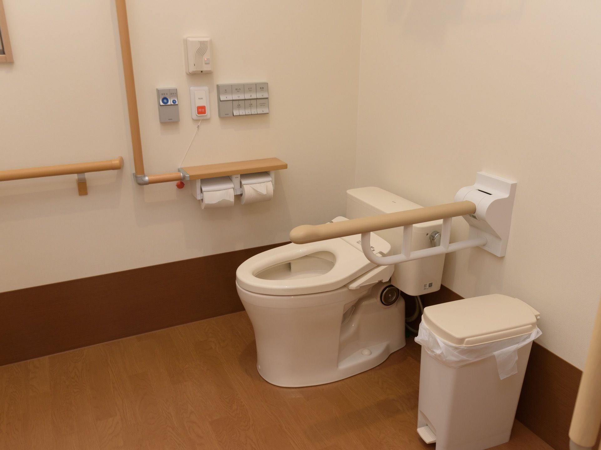 便座ふたのない洋式トイレがあり、白いべダルが付いたゴミ箱が置いてある。壁に手すりや２連のペーパーホルダーがーがある。