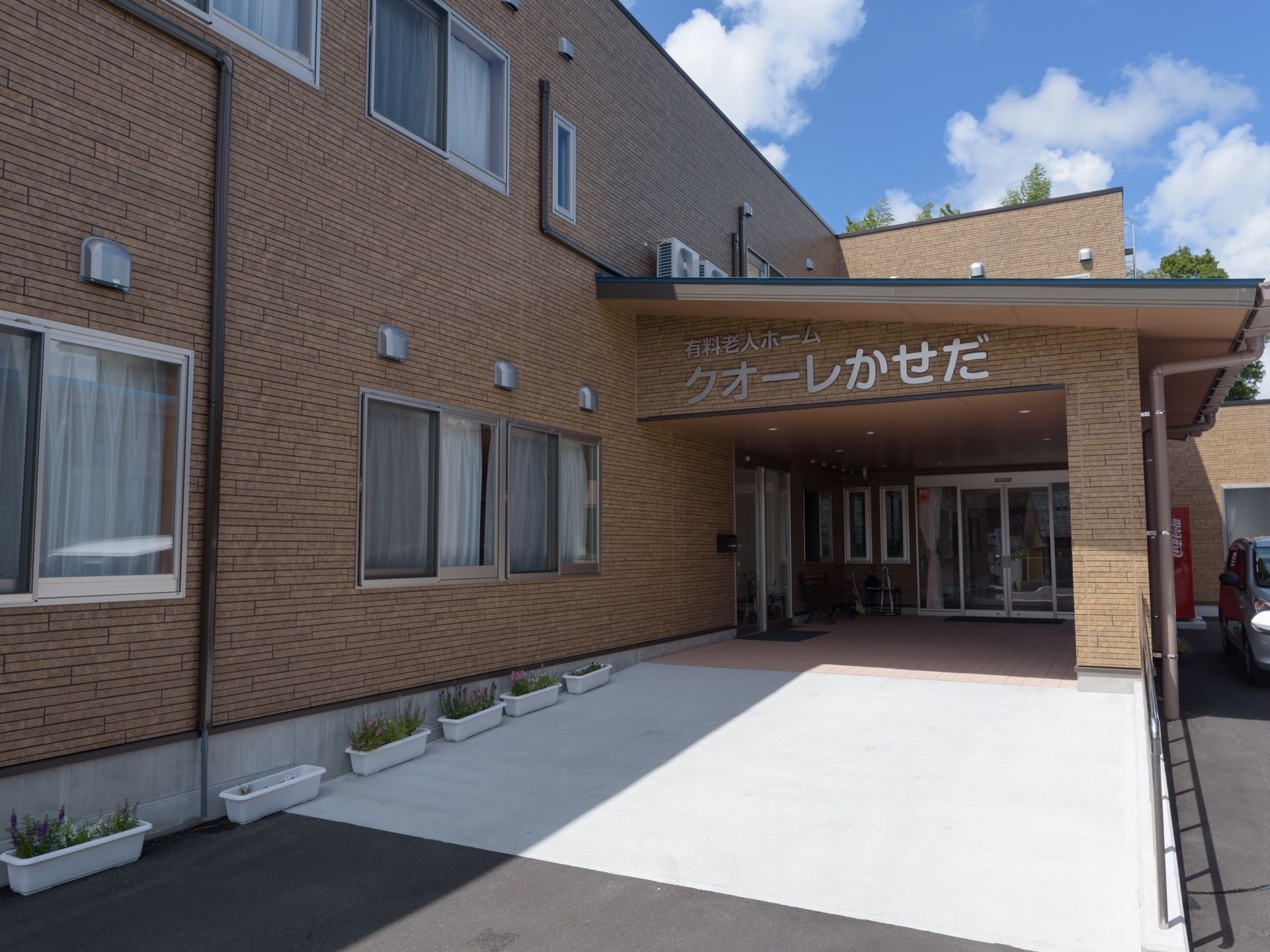 茶色い外壁に銀色の文字で施設の名称が表示されている。スロープには手すりが設置され、プランターが置いてある。