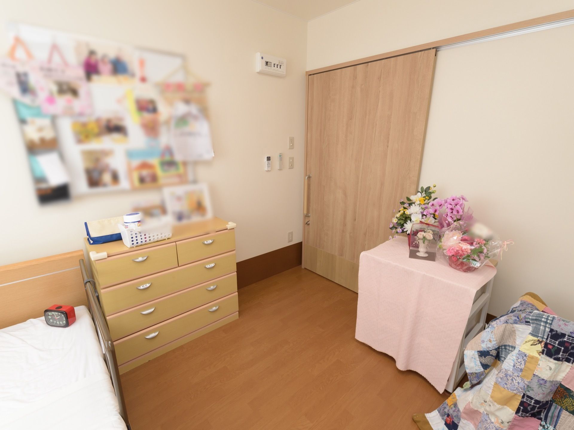 ピンク色の布が掛けられた収納タンスの上にたくさんの花が飾られている。部屋の入り口はスライドドアになっている。