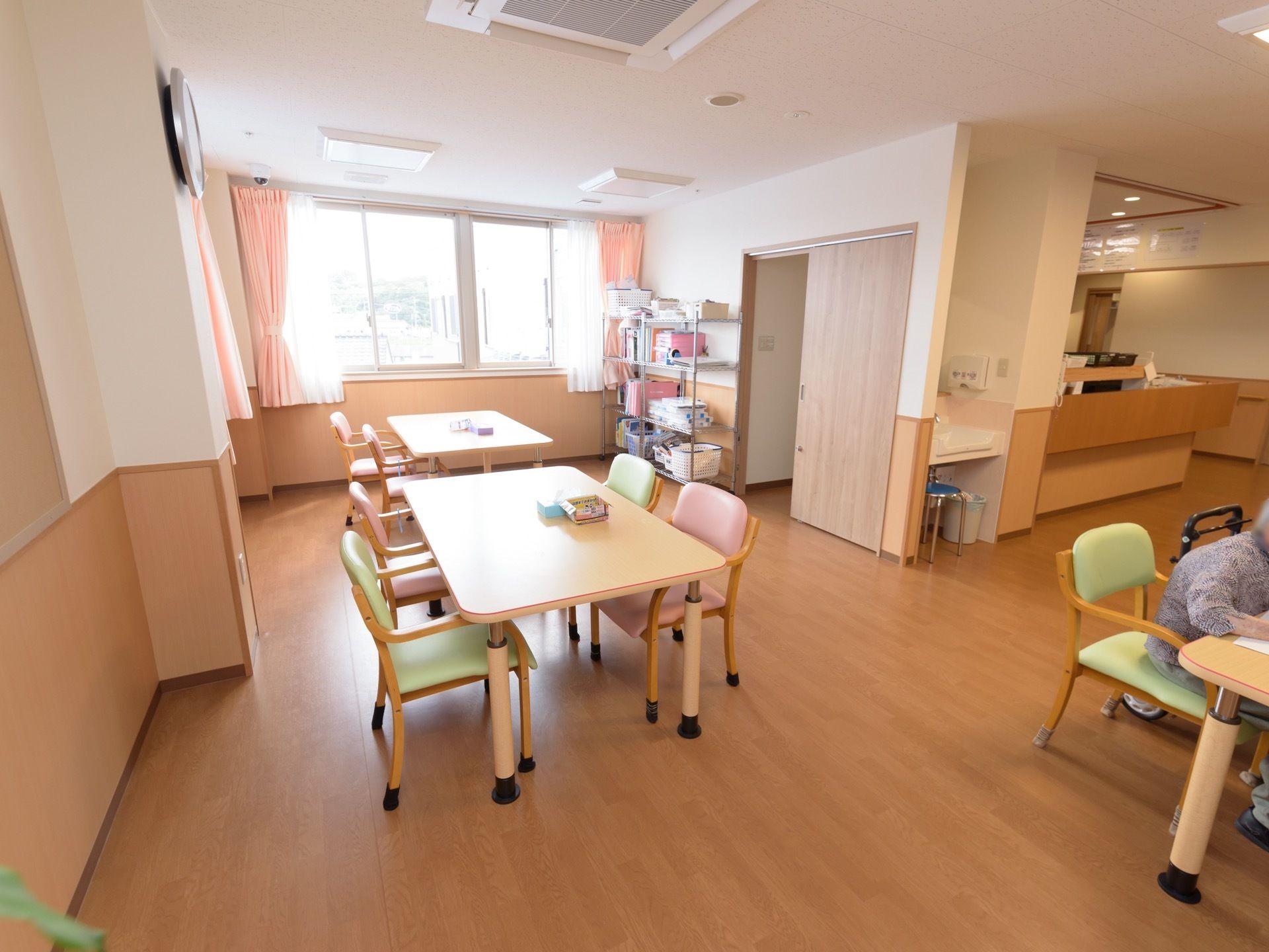 ベージュ色と白色を基調とした部屋に、ダイニングテーブルや椅子が置いてある。サーモンピンクのカーテンが掛けられた腰高窓がある。