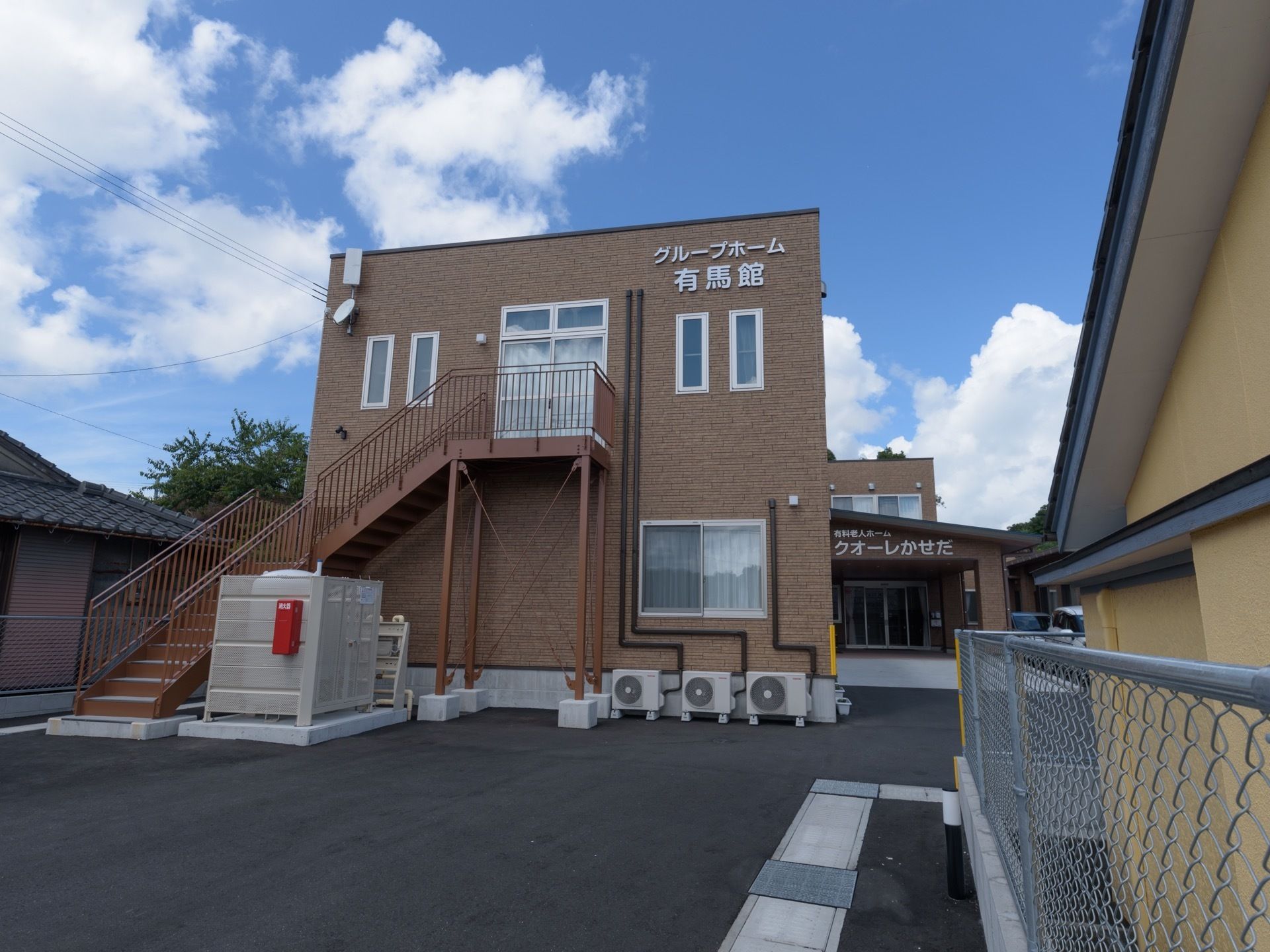 アスファルトで平らに舗装された敷地に2階建ての住宅が建っている。外壁は茶色いタイルで装飾されている。